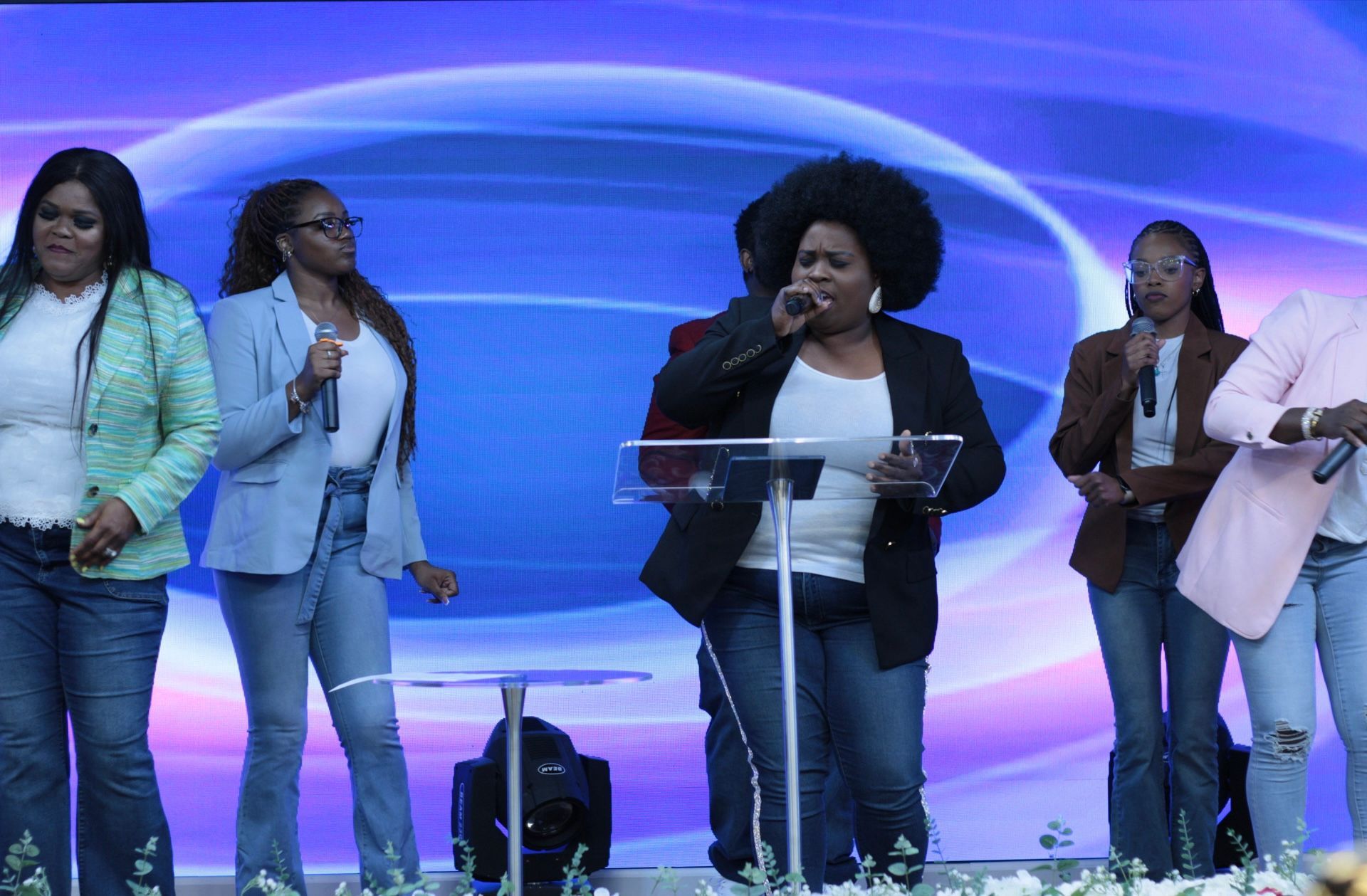 Women singing into microphones on a stage with a blue background; wearing jeans and jackets.