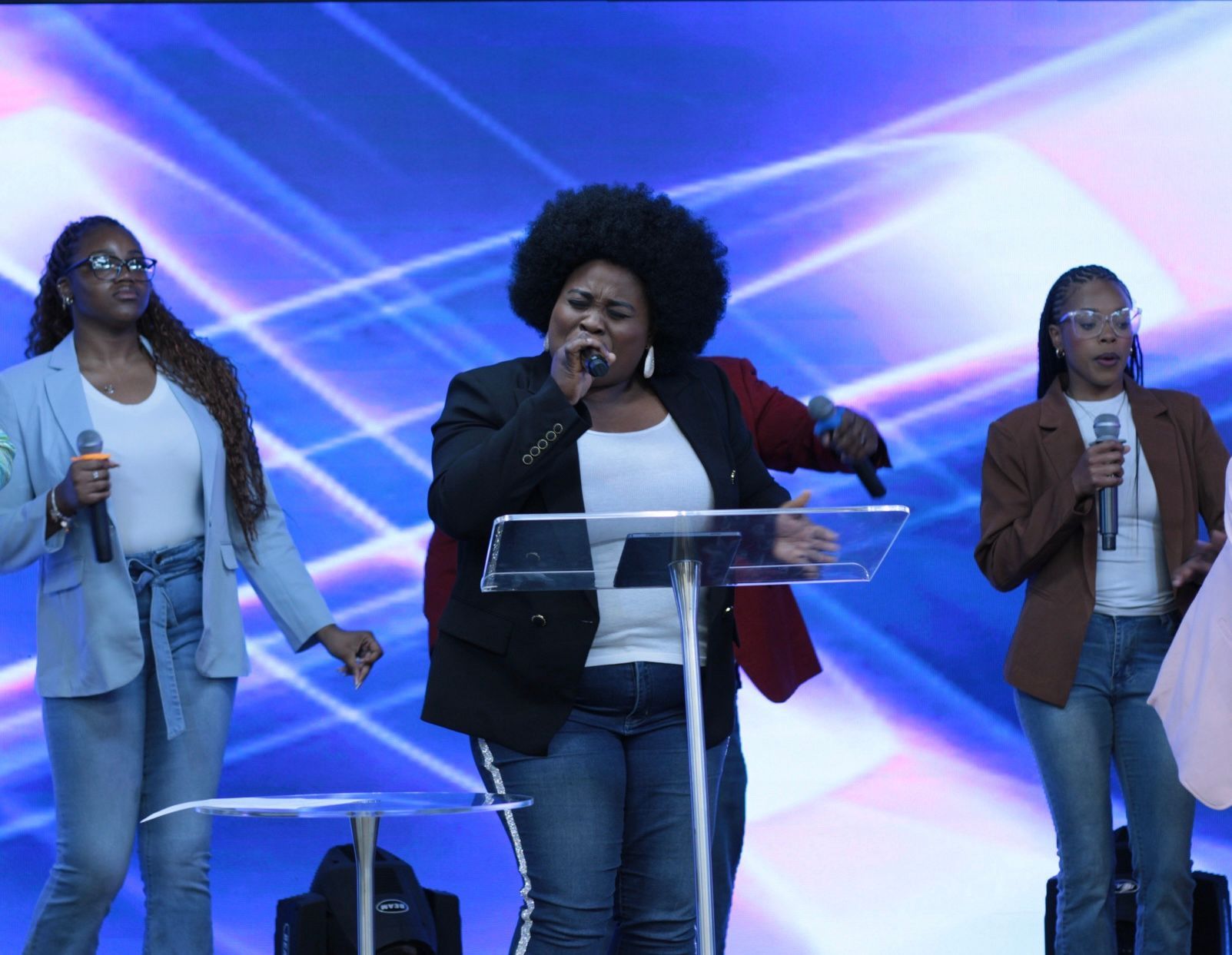 A gospel choir singing on stage, three Black women with microphones, blue backdrop.