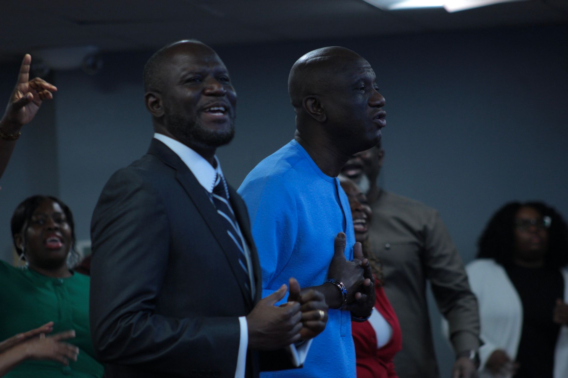 Two Black men singing in a group, one in a suit, one in blue, others in the background.