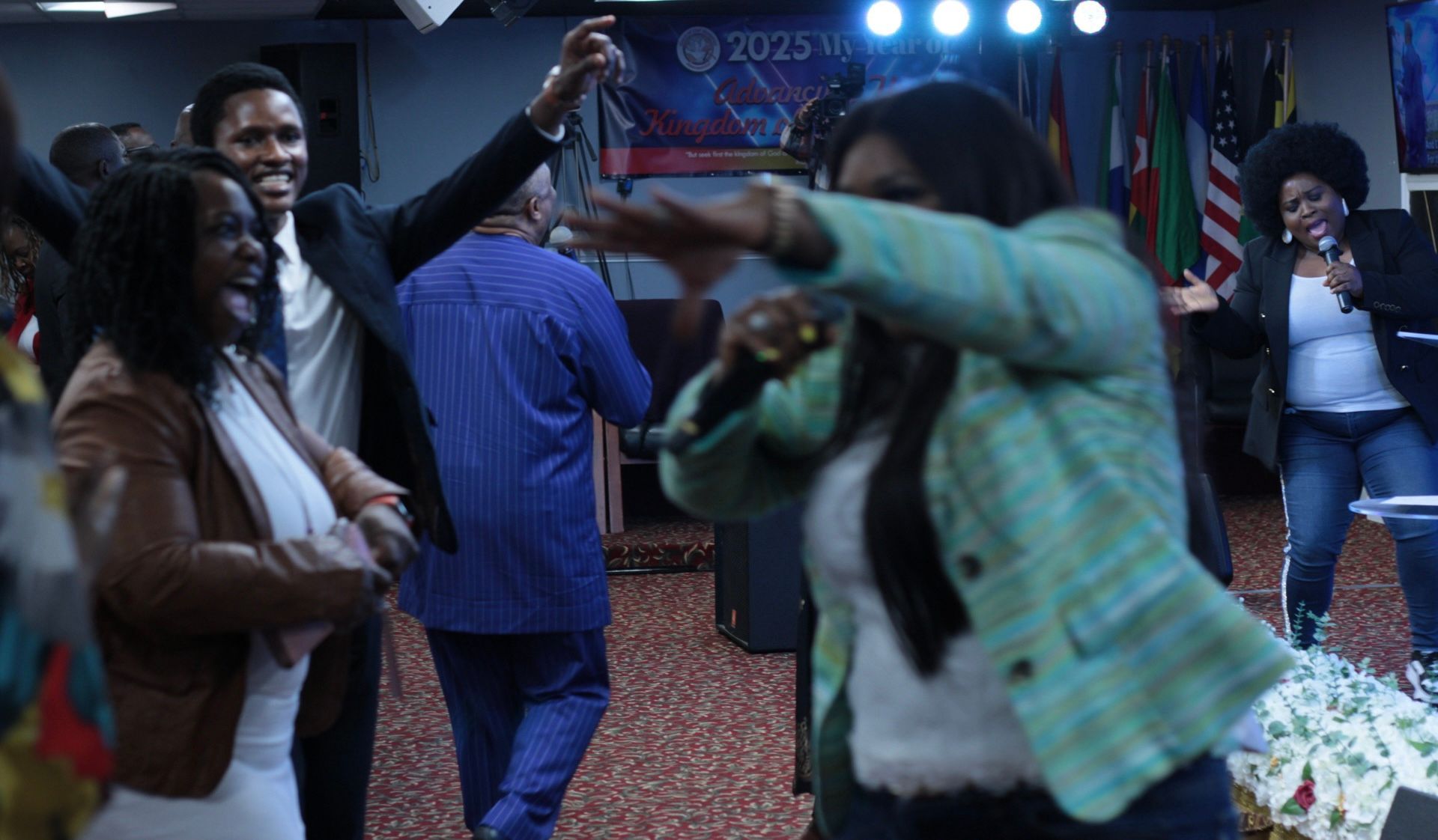 People dancing joyfully in a brightly lit room, with a woman singing on a stage, flags in the background.