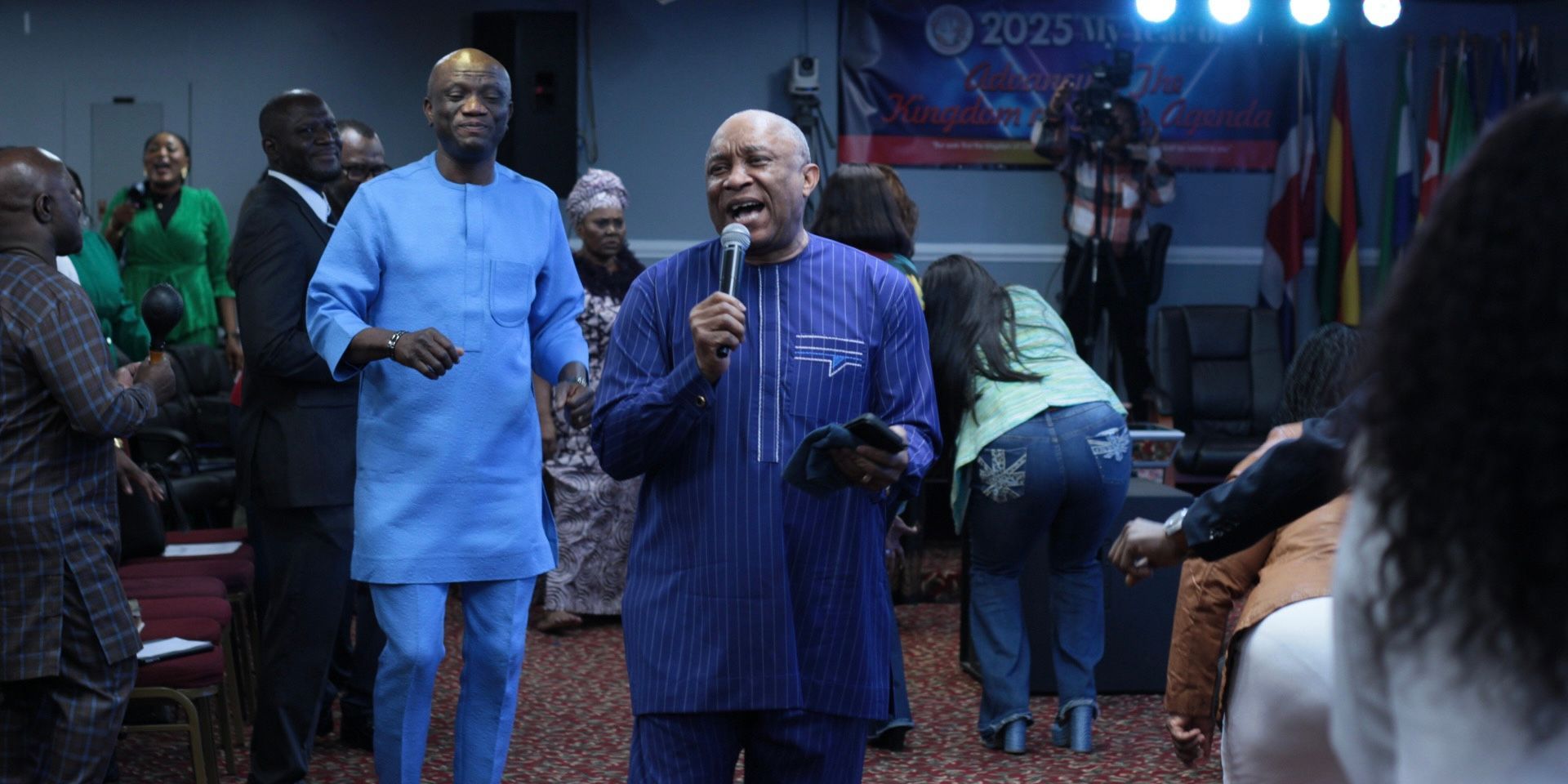 Man speaking into a microphone, surrounded by people in a brightly lit room.