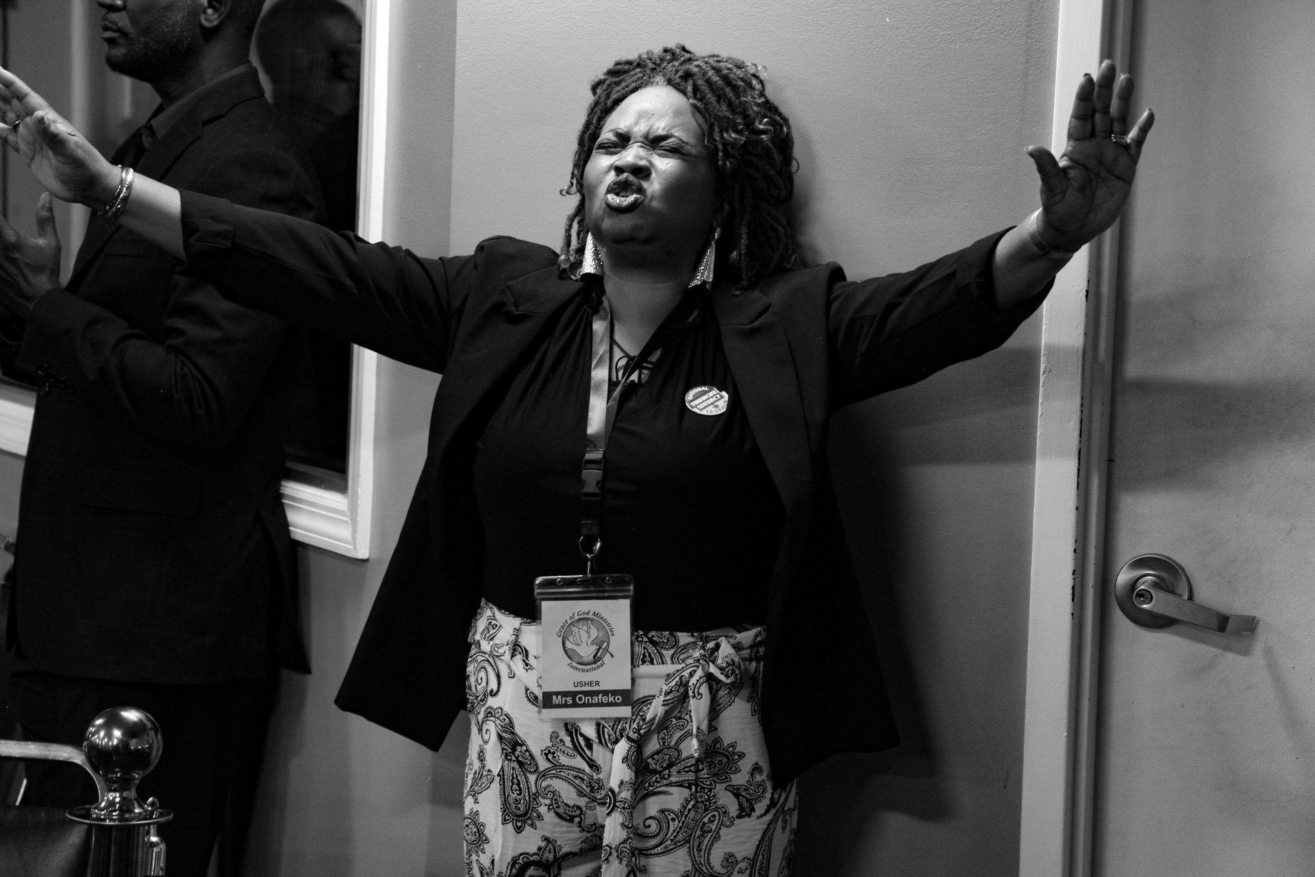 Woman with arms raised, eyes closed, wearing a blazer and floral pants, near a door.