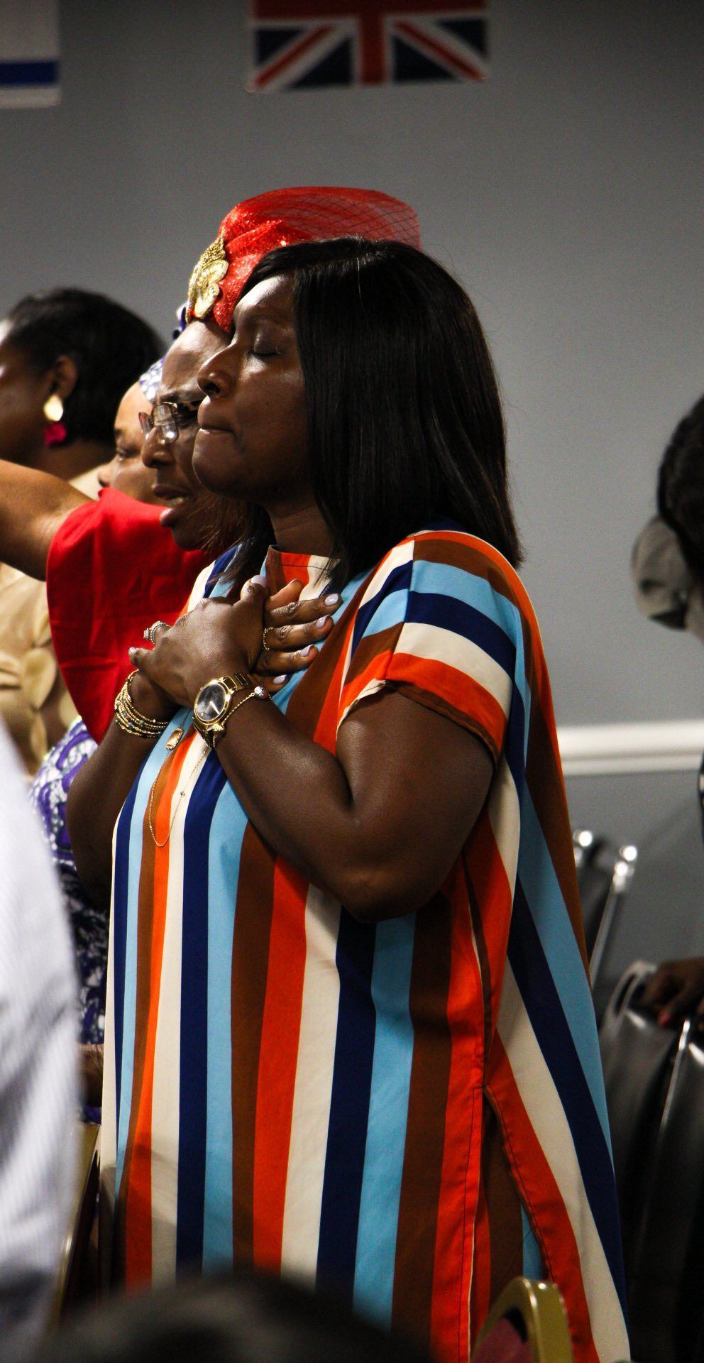 Woman in striped dress with hands on chest, eyes closed, in a room with others.