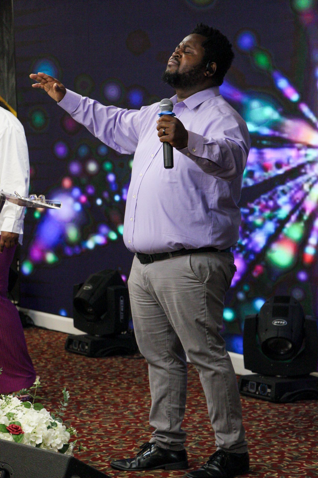 Man singing on stage with arms raised, wearing a purple shirt and gray pants.