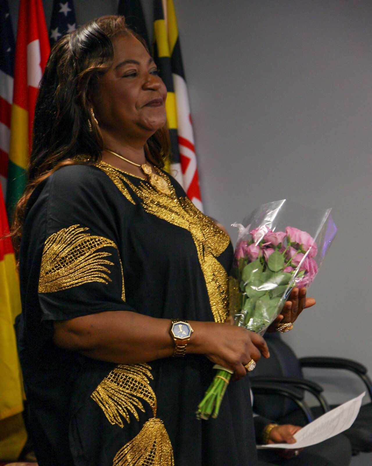 Woman in black dress with gold embroidery holding flowers, smiling at a podium with flags in the background.
