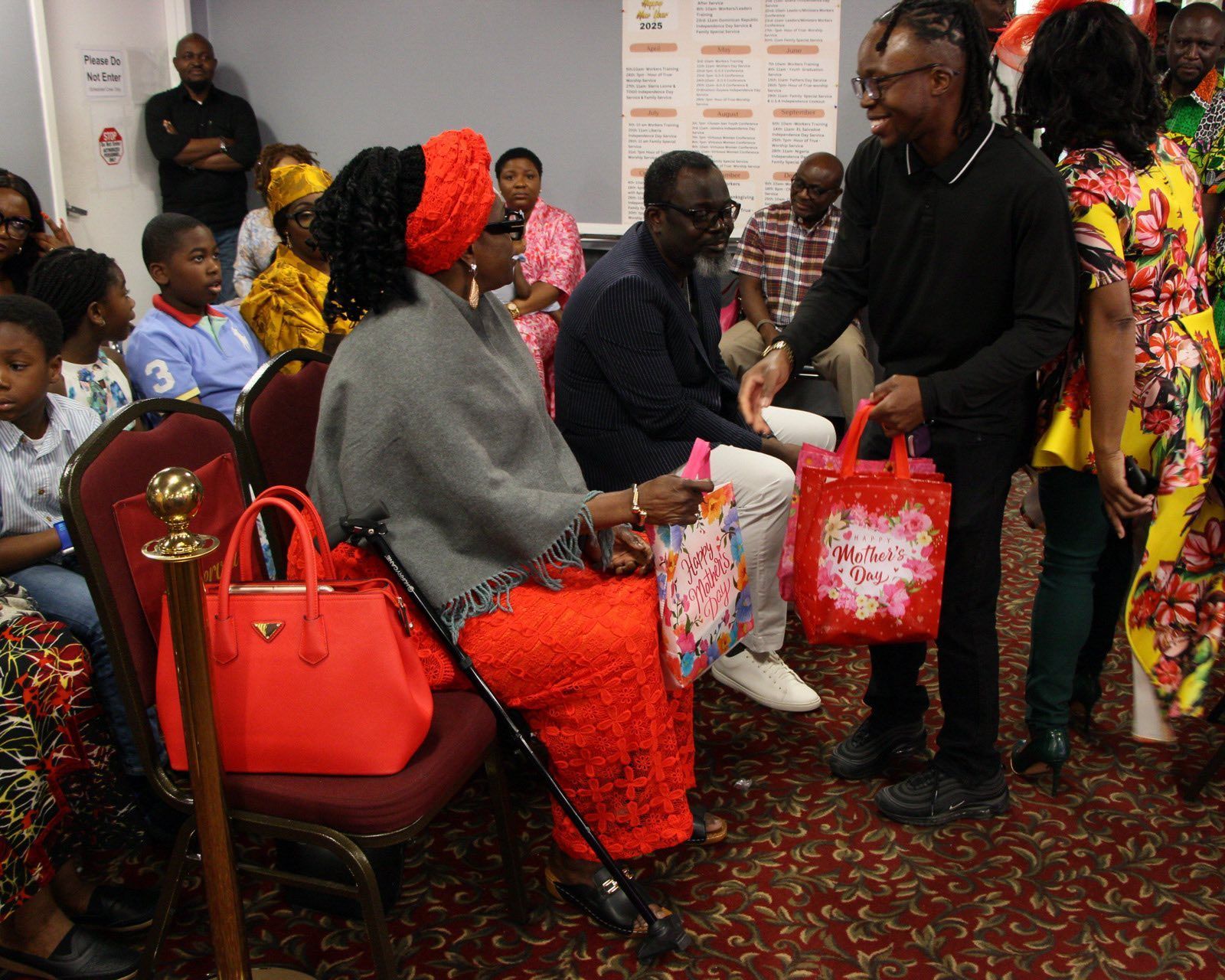 Person giving a gift bag to an elderly woman, likely at an event. Red and gold colors are prominent.
