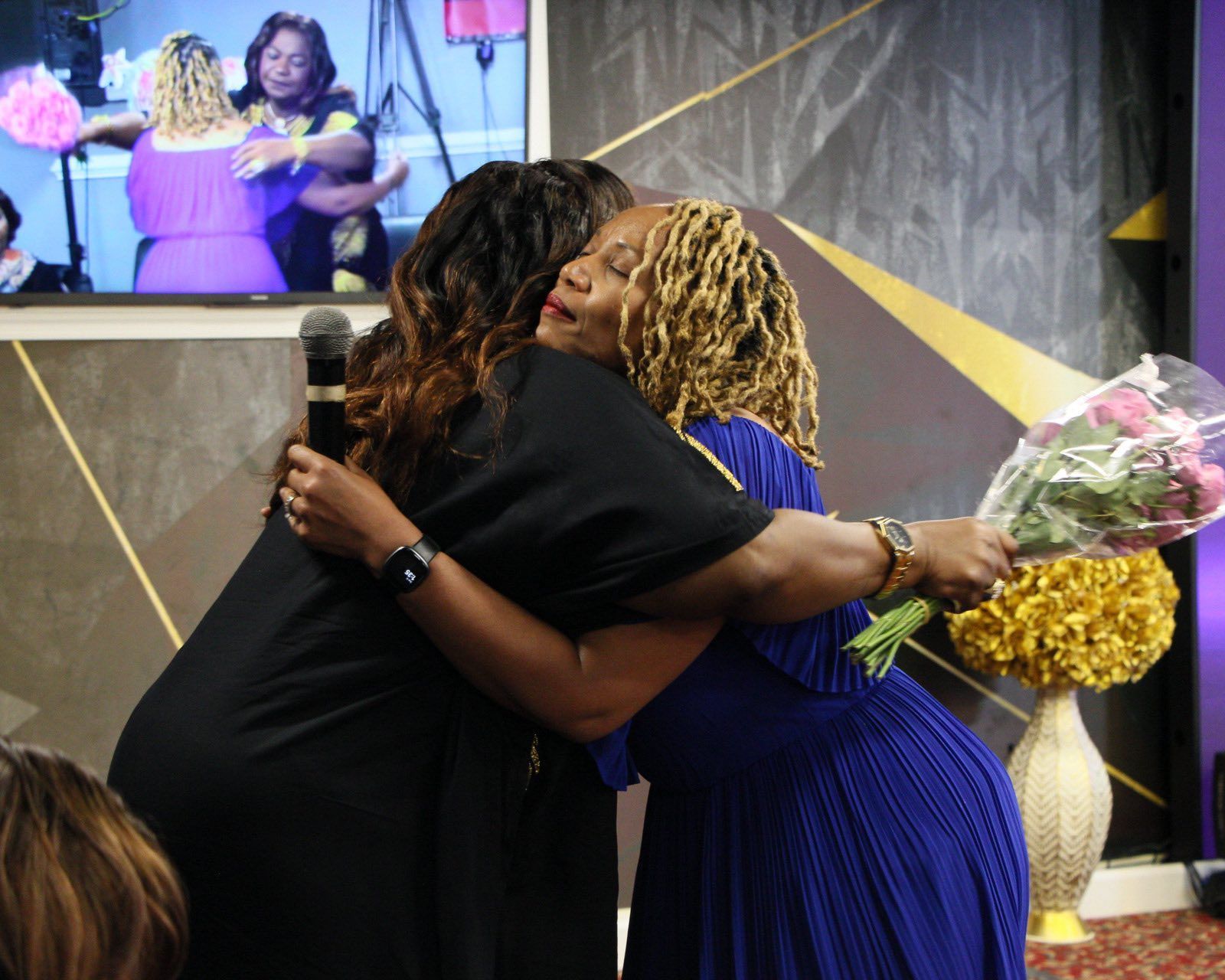 Two women embrace, one holding roses. They are in a brightly lit setting with a screen showing others hugging.