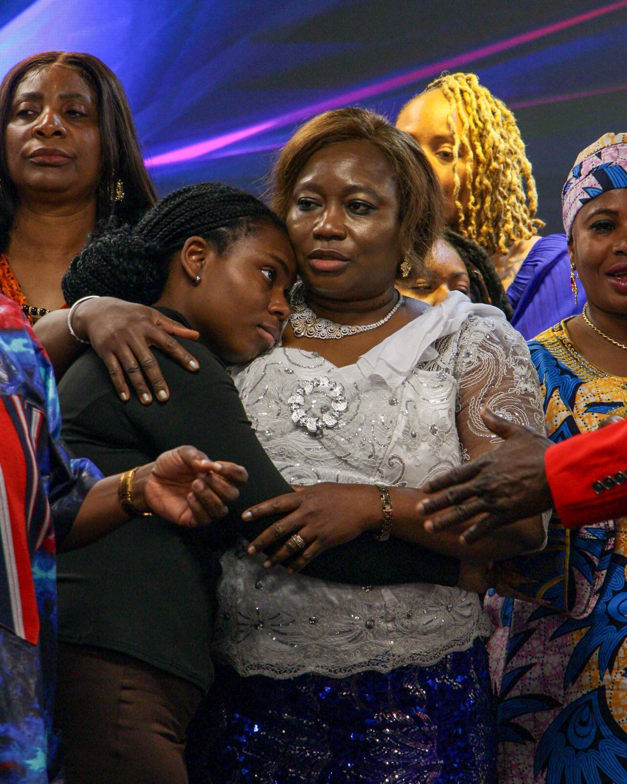 Woman in white lace gown hugs a girl, surrounded by other women in colorful clothing.