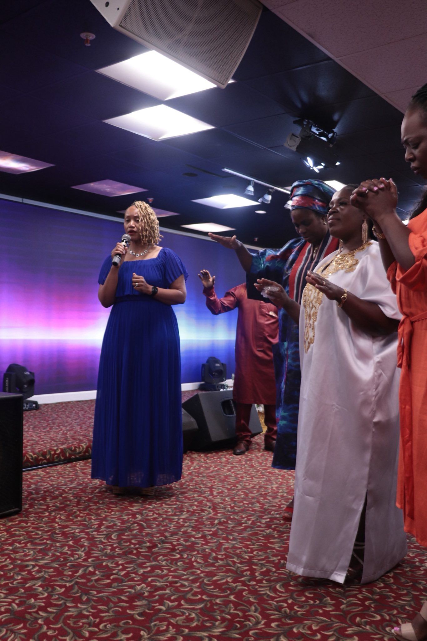 Woman in blue dress singing; others raise hands, singing, on stage with red carpet and screens.
