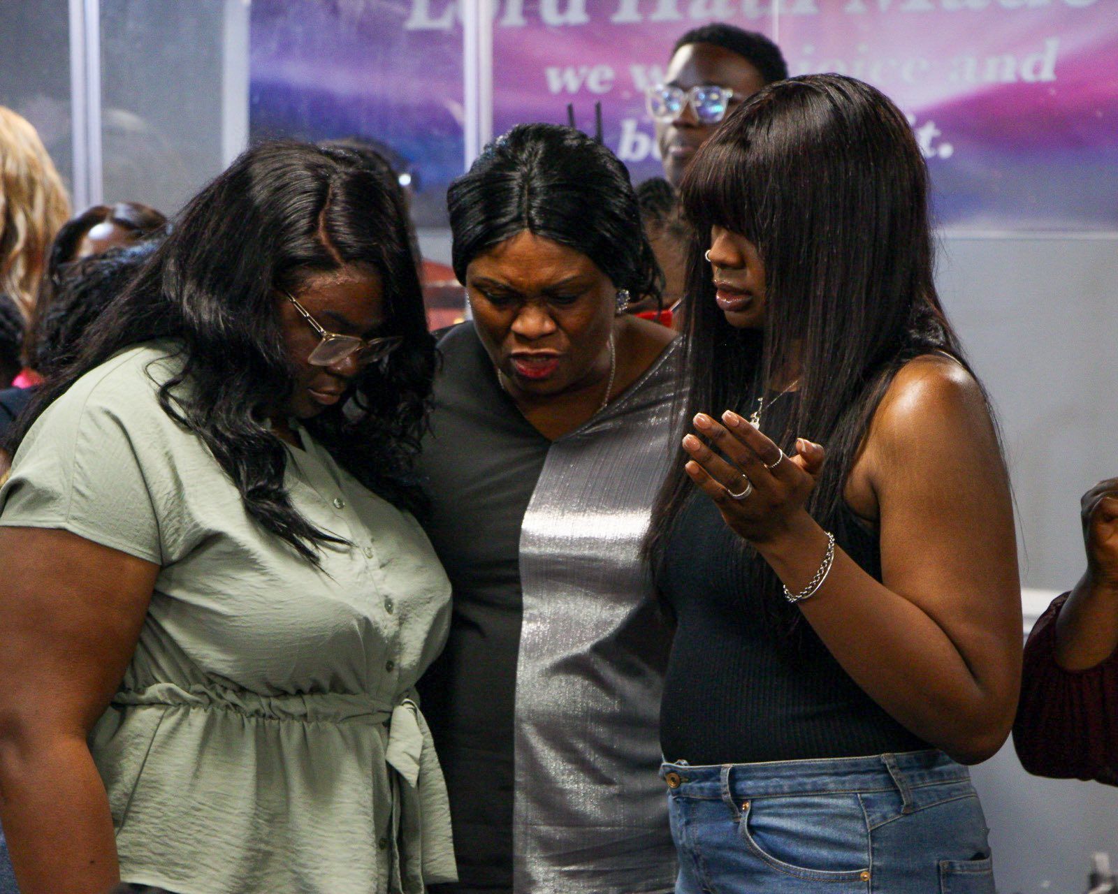 Three women praying in a church, heads bowed, faces concerned. Soft lighting, purple background.