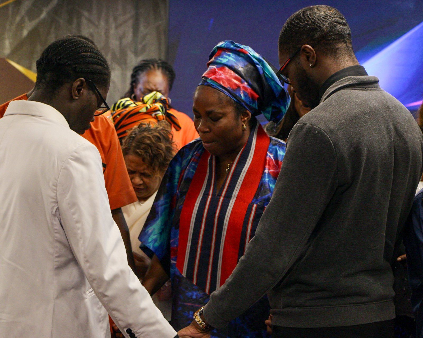 People praying together in a church. Several are touching hands, some wearing colorful African attire.