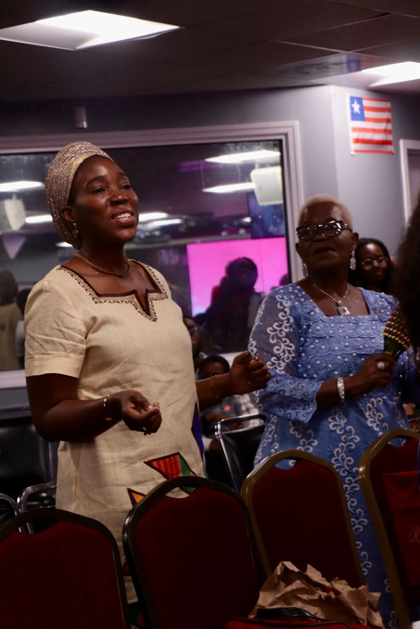 Two women singing in a room with a Liberian flag, one wearing cream, the other in blue.