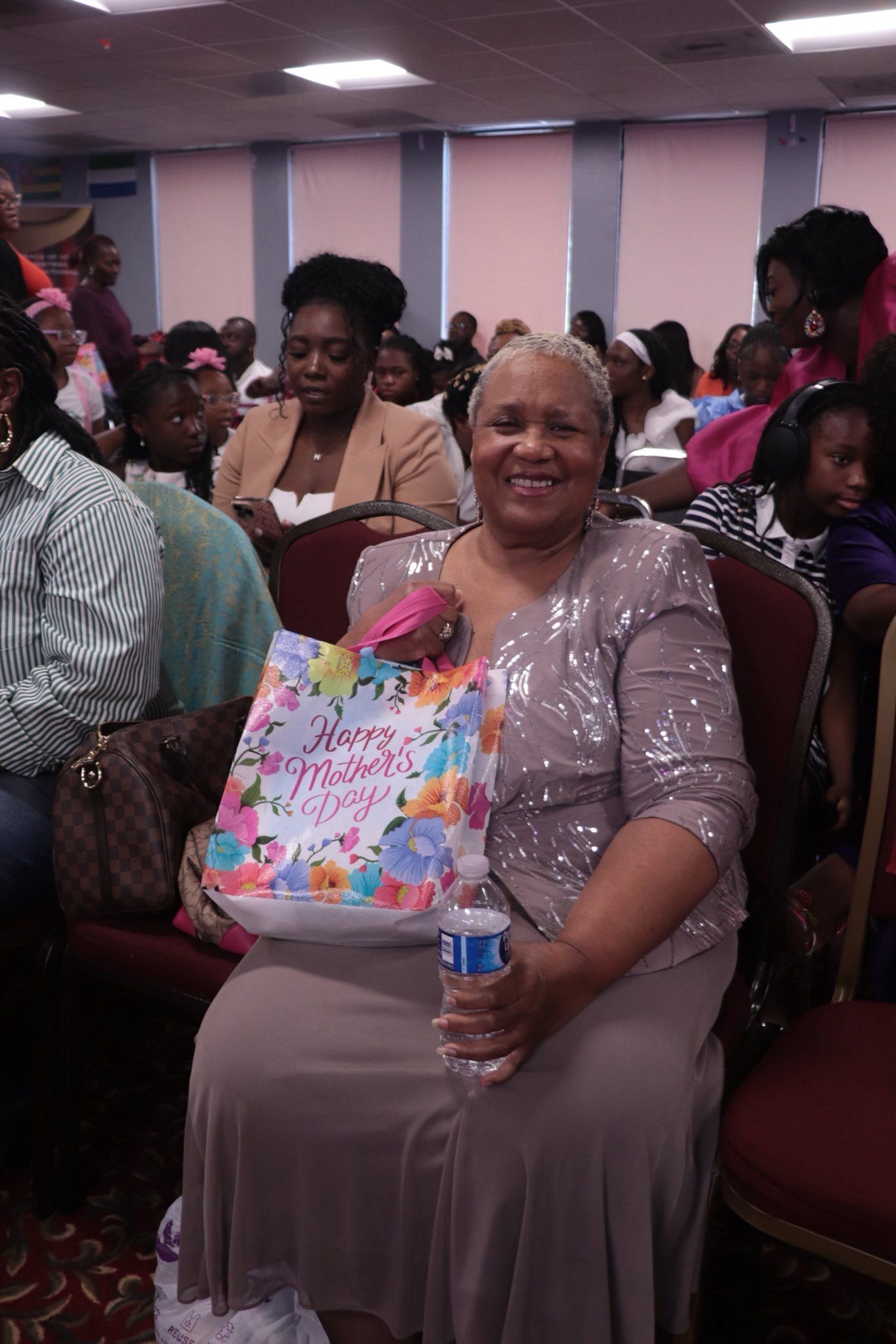 Woman in a sparkly top and skirt smiles holding a gift bag. Sitting in a room with people.