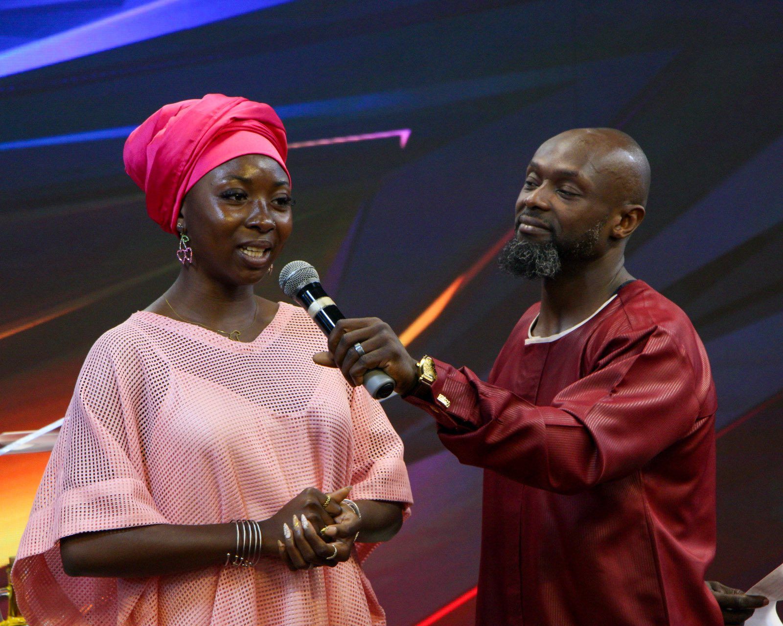 Woman in pink speaks into a mic as a man in red holds it. They stand on a stage.