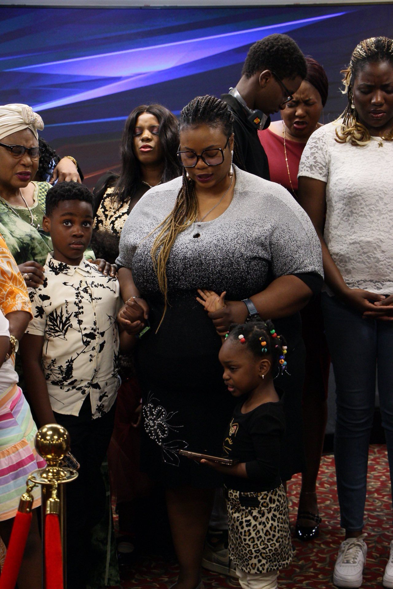 A diverse group of people praying together. Focus on a woman, child, and boy holding hands, near a velvet rope.