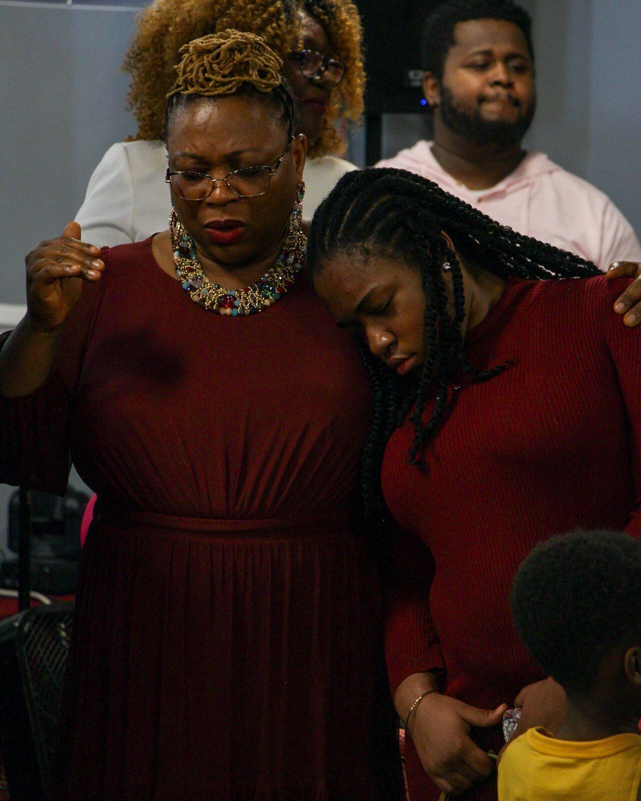People in a church, praying. Woman in maroon dress with hand raised, young woman leaning on her, all eyes closed.