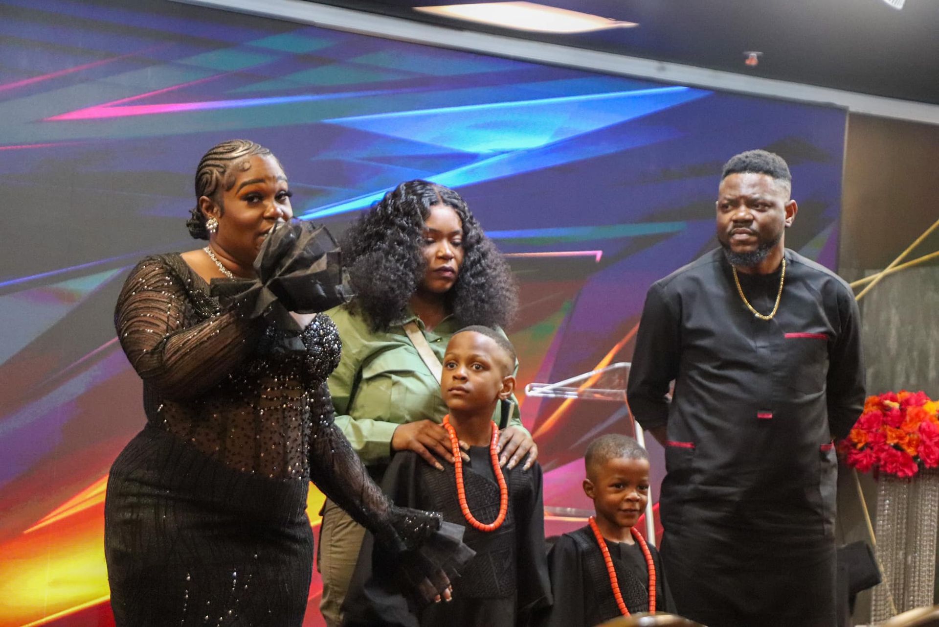 Family standing onstage: woman in black dress speaks, flanked by others, with two children in traditional outfits.