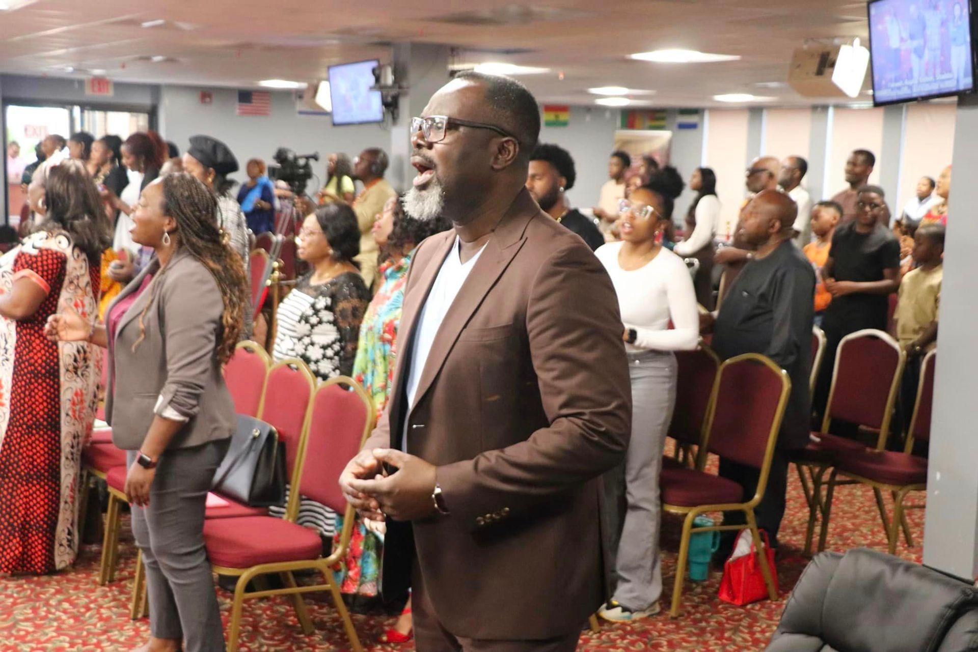 A diverse group of people singing with hands raised in a church; brown suit, red chairs.