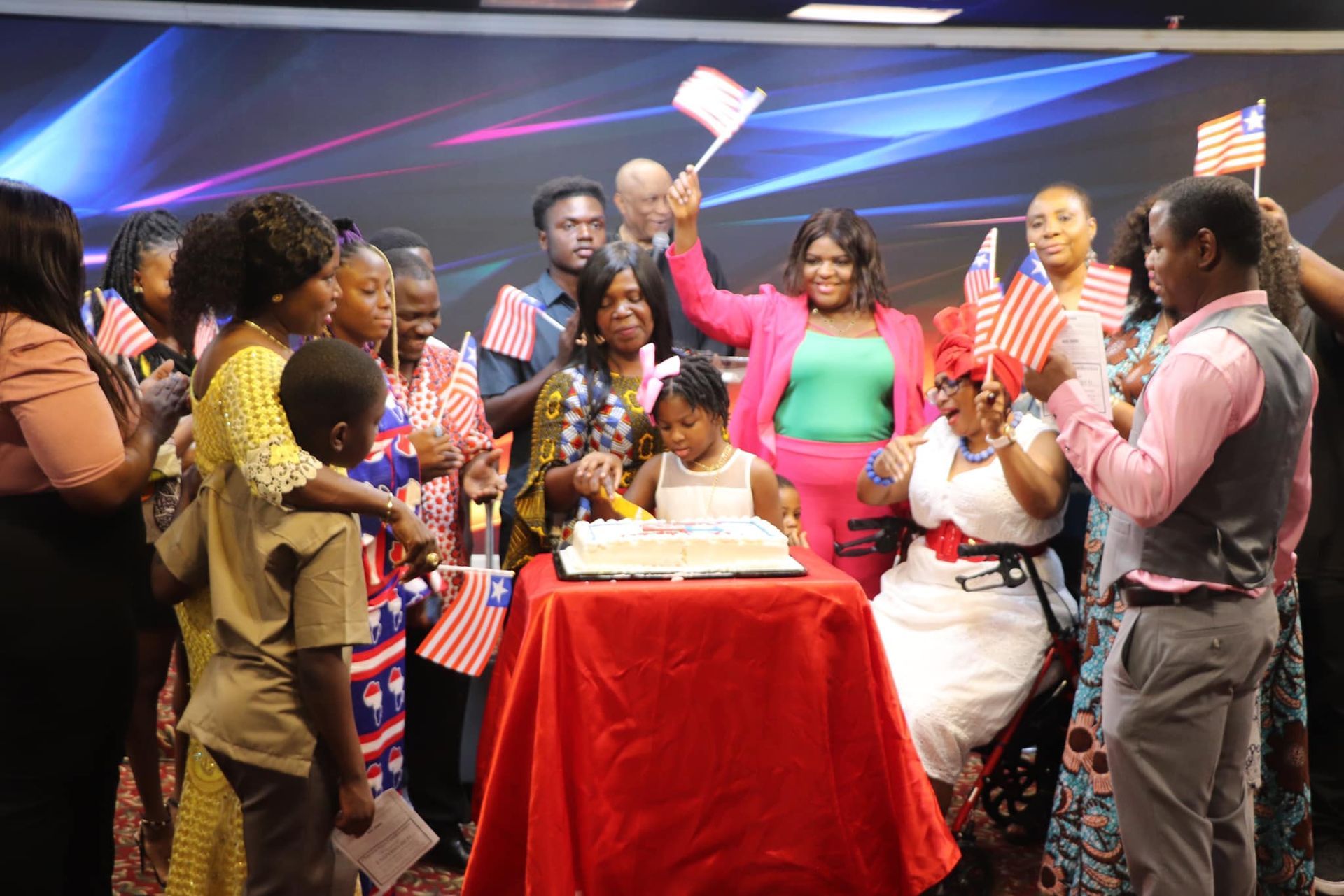 Group celebrating with flags and cake. People are gathered around a table, with a woman in pink waving a flag.