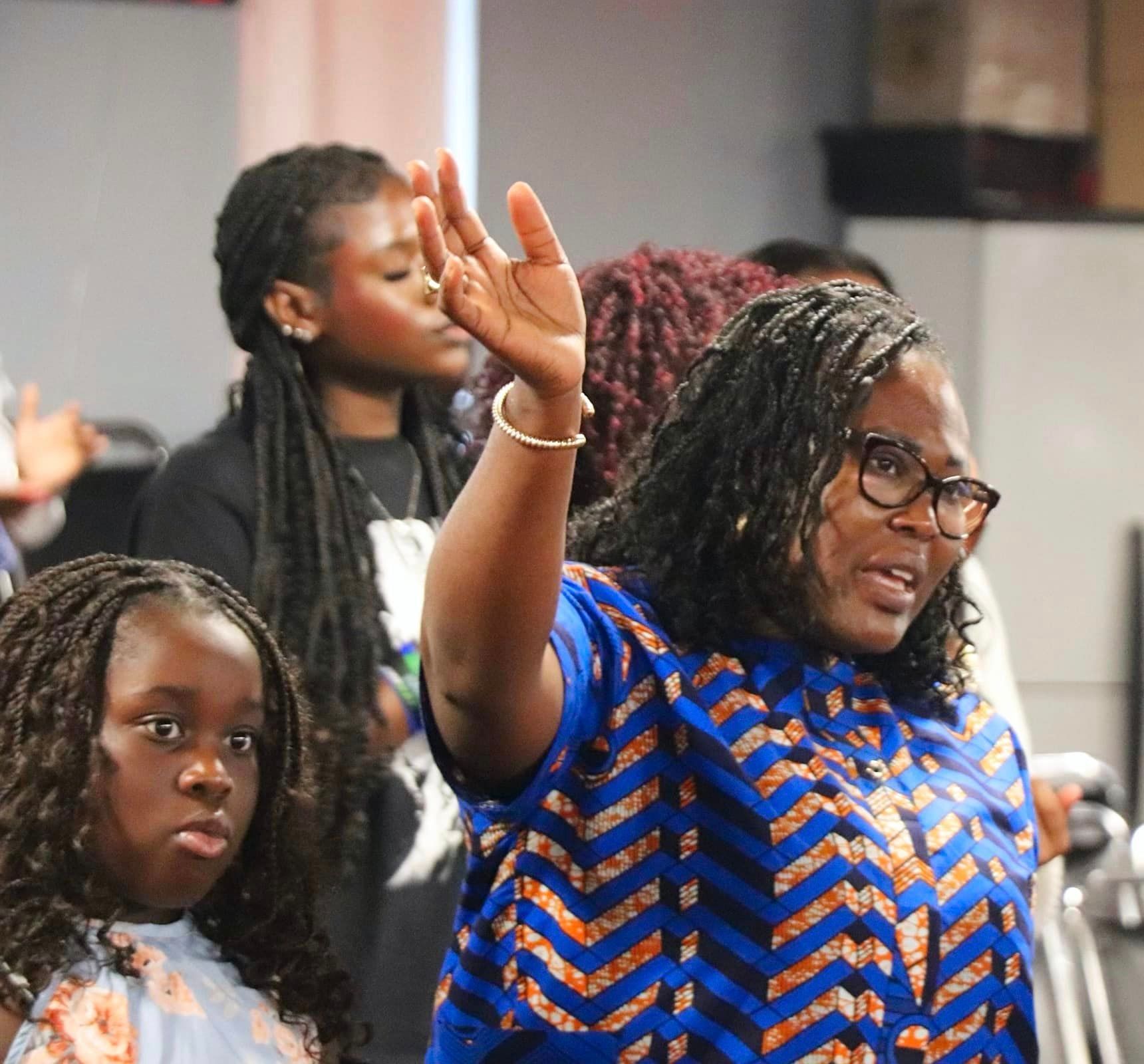 Woman with glasses raises hand in worship, others around her. Blue and gold patterned shirt. Indoors.
