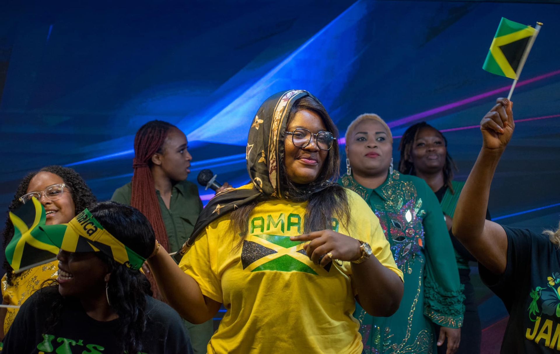 Group of Jamaican women celebrating, waving flags. Woman in yellow shirt points. Blue background.