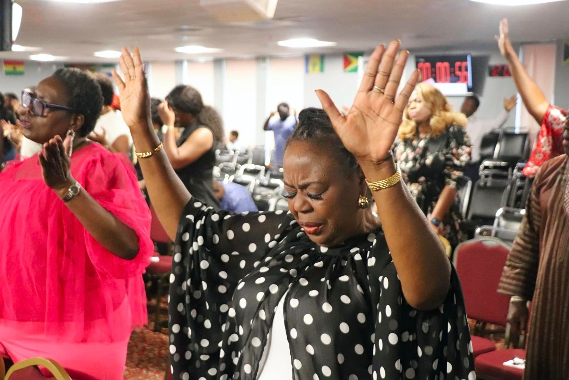 People raising hands in worship, in a brightly lit room, one woman in black and white polka dots.