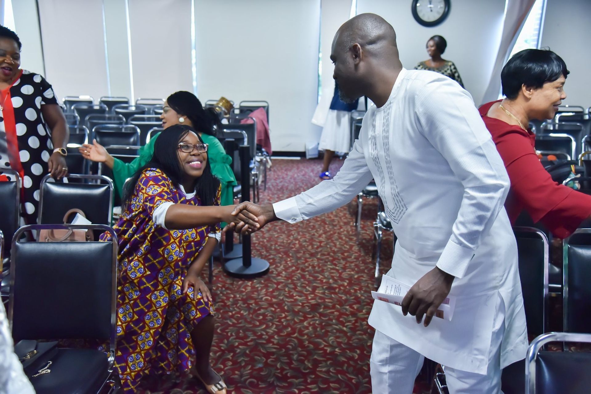 People in a room: man in white shaking hands with a woman in patterned dress, smiling. Other people around.