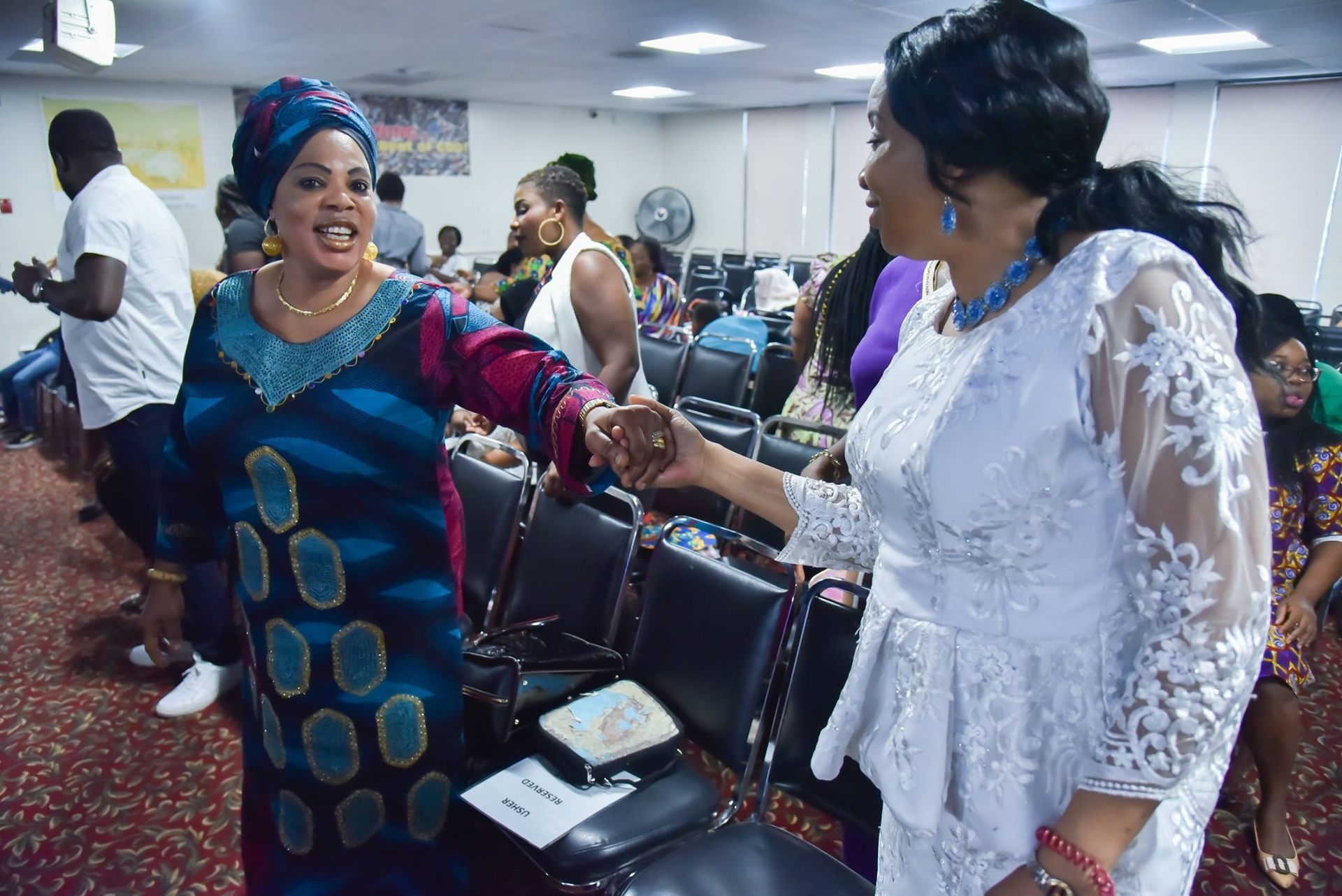 Two women in colorful African attire greet each other in a crowded room, smiling and shaking hands.