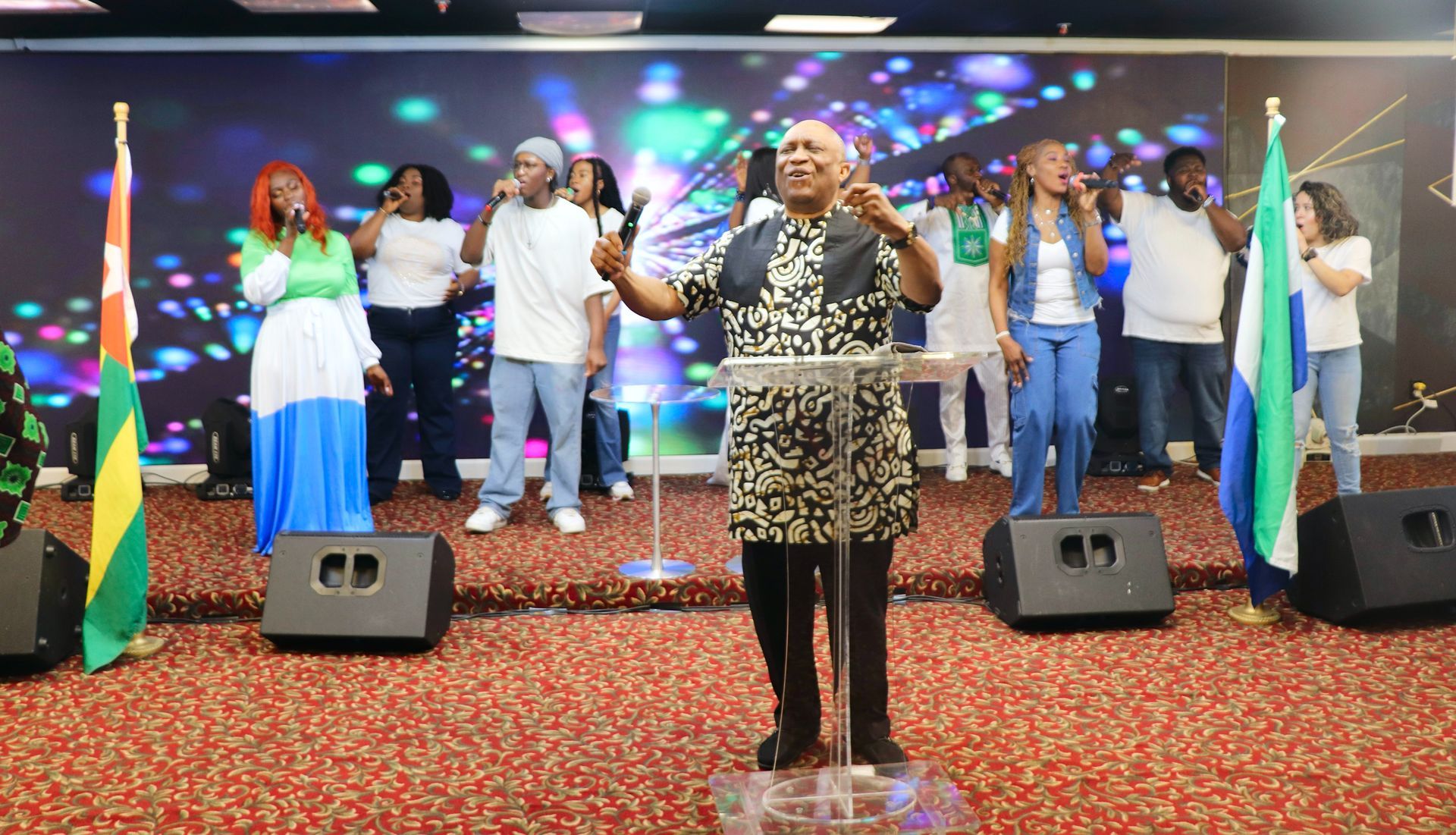 Choir singing on stage with flags; man in patterned shirt leads with arms raised.