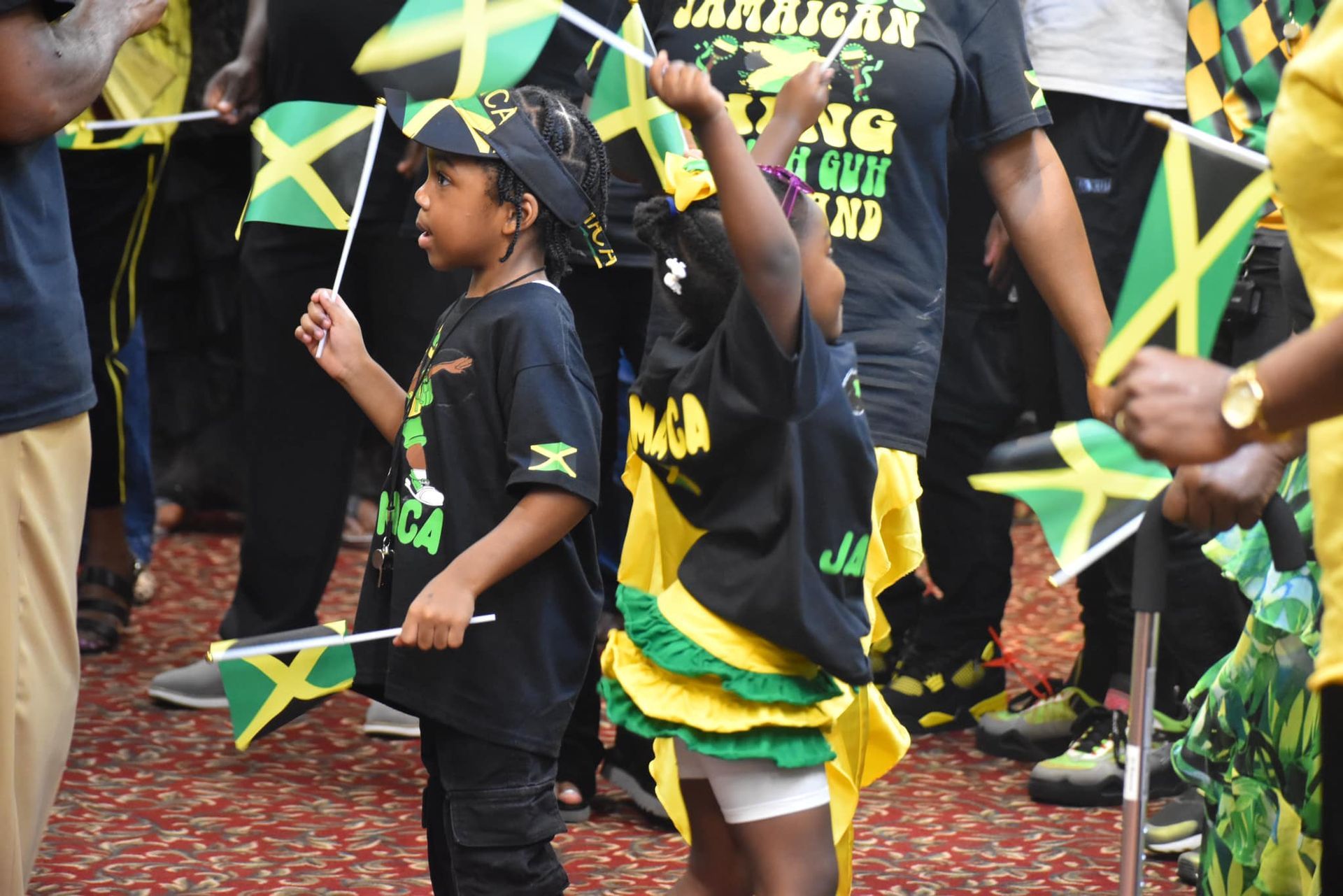 Two children waving Jamaican flags, wearing black and yellow outfits, in a crowd.