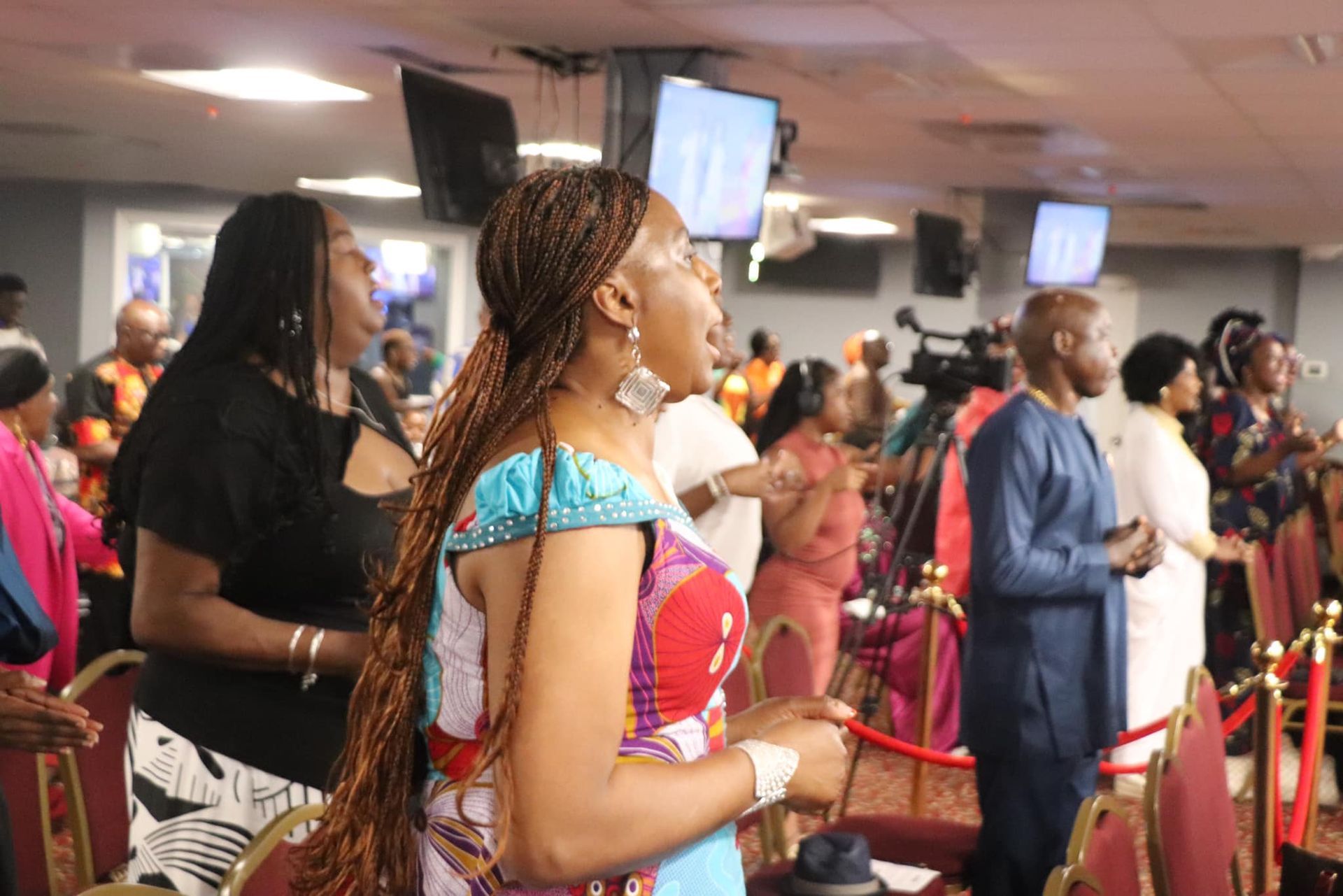 People in a church service, singing and raising hands. Colorful clothing, warm tones, and overhead screens.