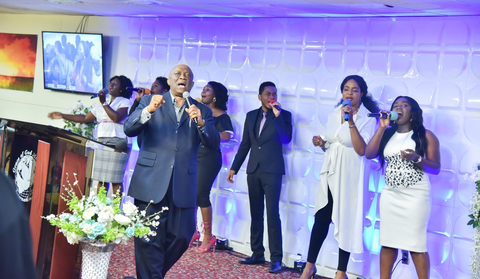Choir singing in a church with blue lighting. Men and women, microphones, singing, and a floral arrangement.