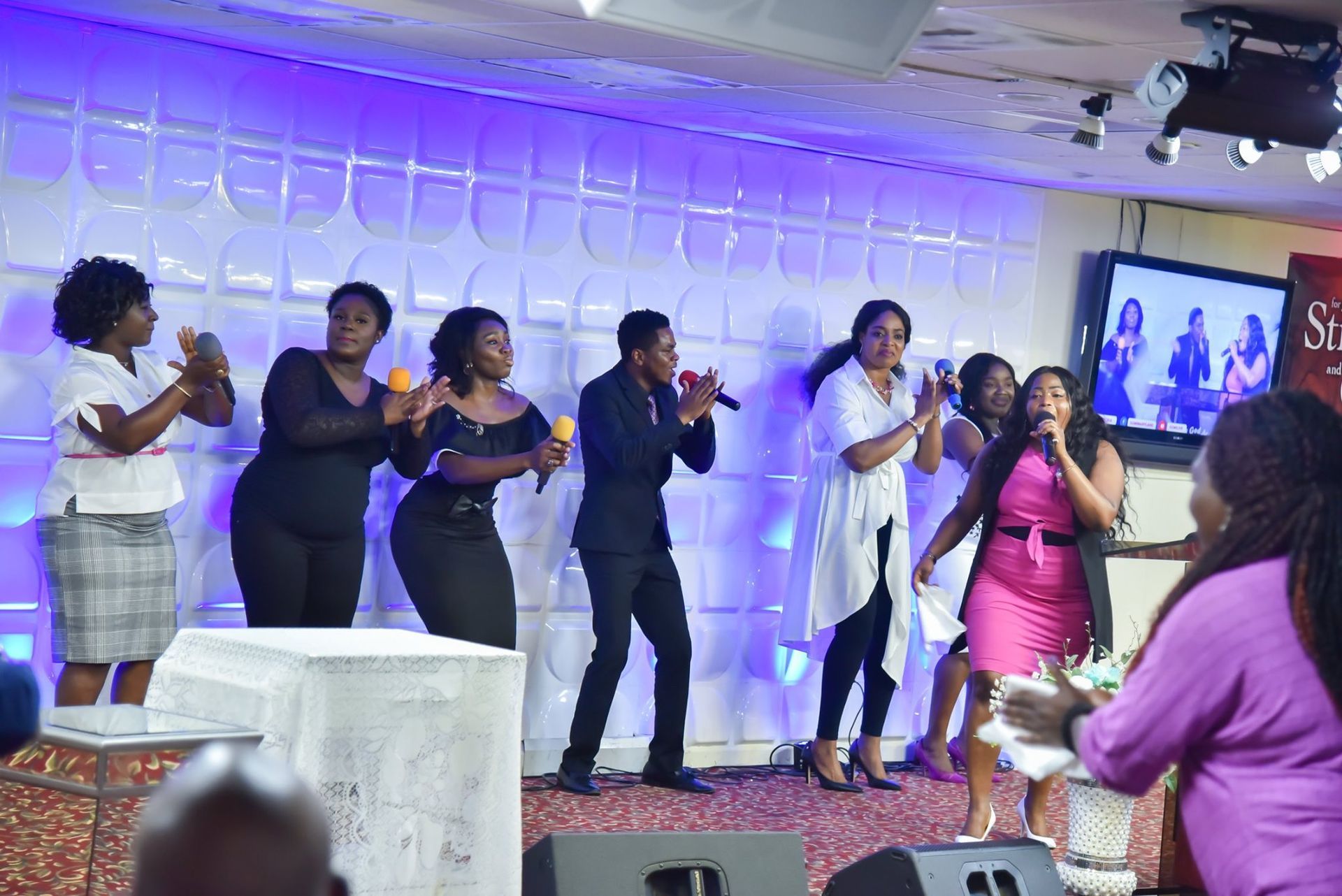 A group singing on a stage with microphones; women and men are clapping.