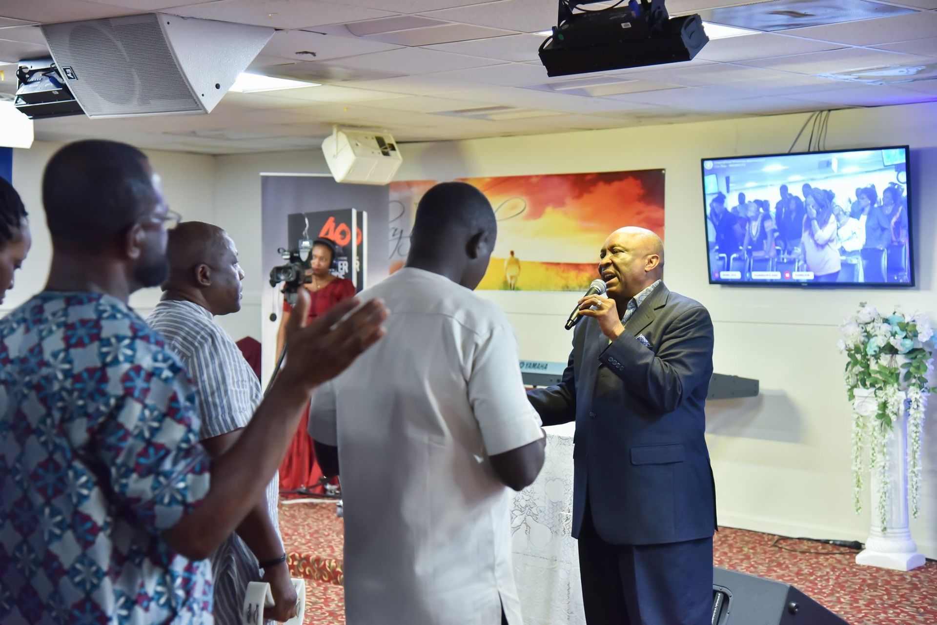 Men in a church listen to a pastor speaking into a microphone. A screen shows a congregation.