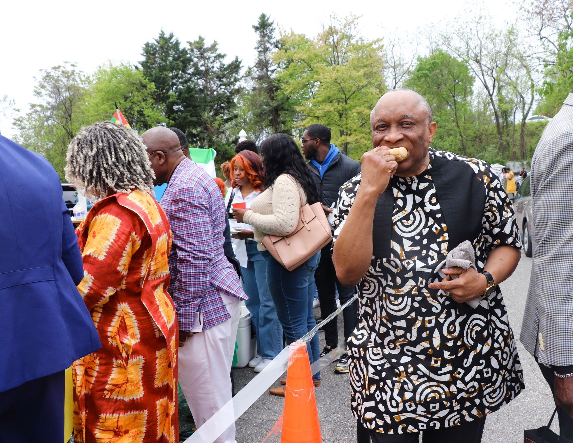 Man eating food, wearing black patterned shirt, at an outdoor event with other people.
