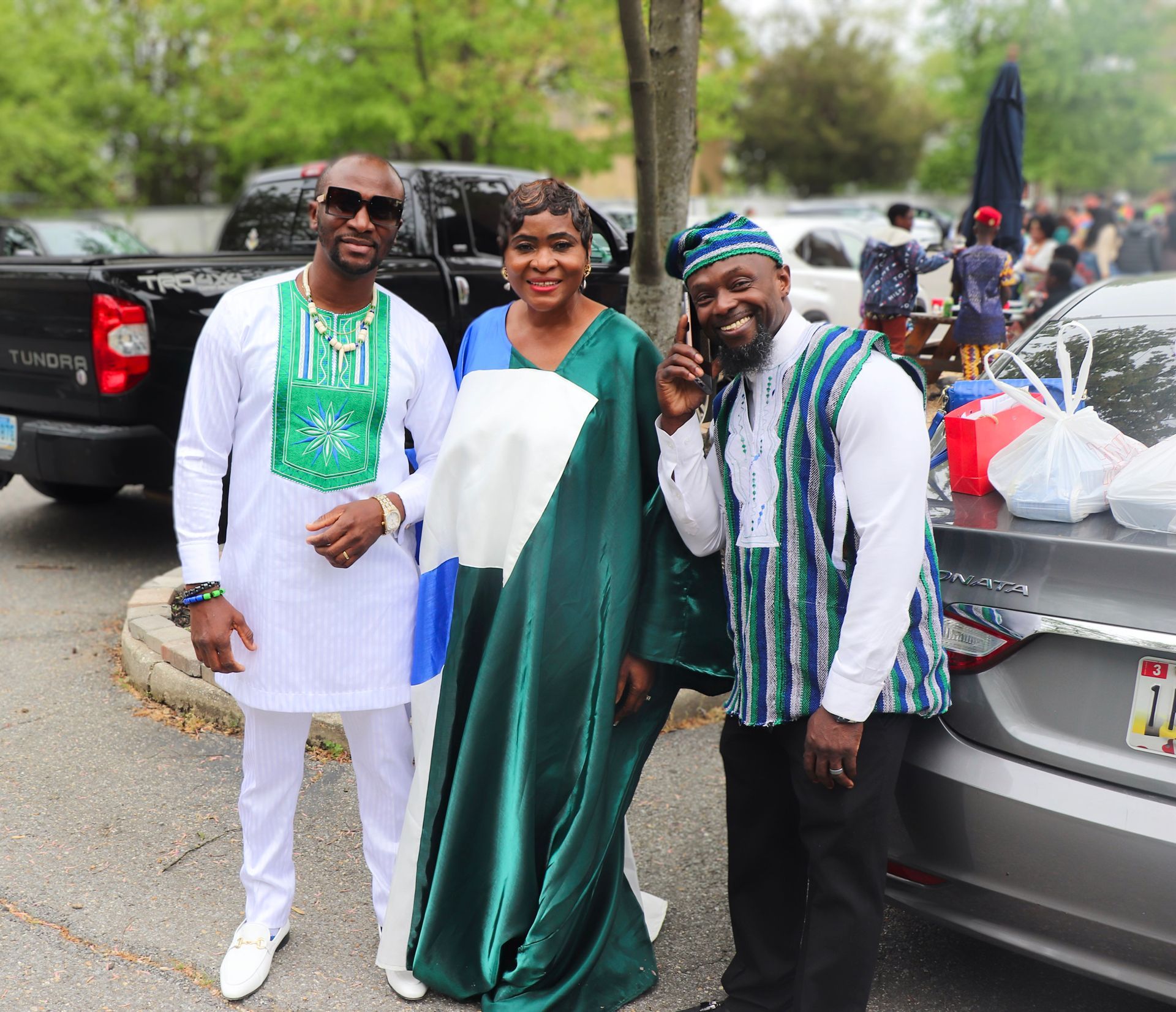 Three people in festive outfits pose outside. Woman in green/blue dress, men in white outfits with green/blue accents.