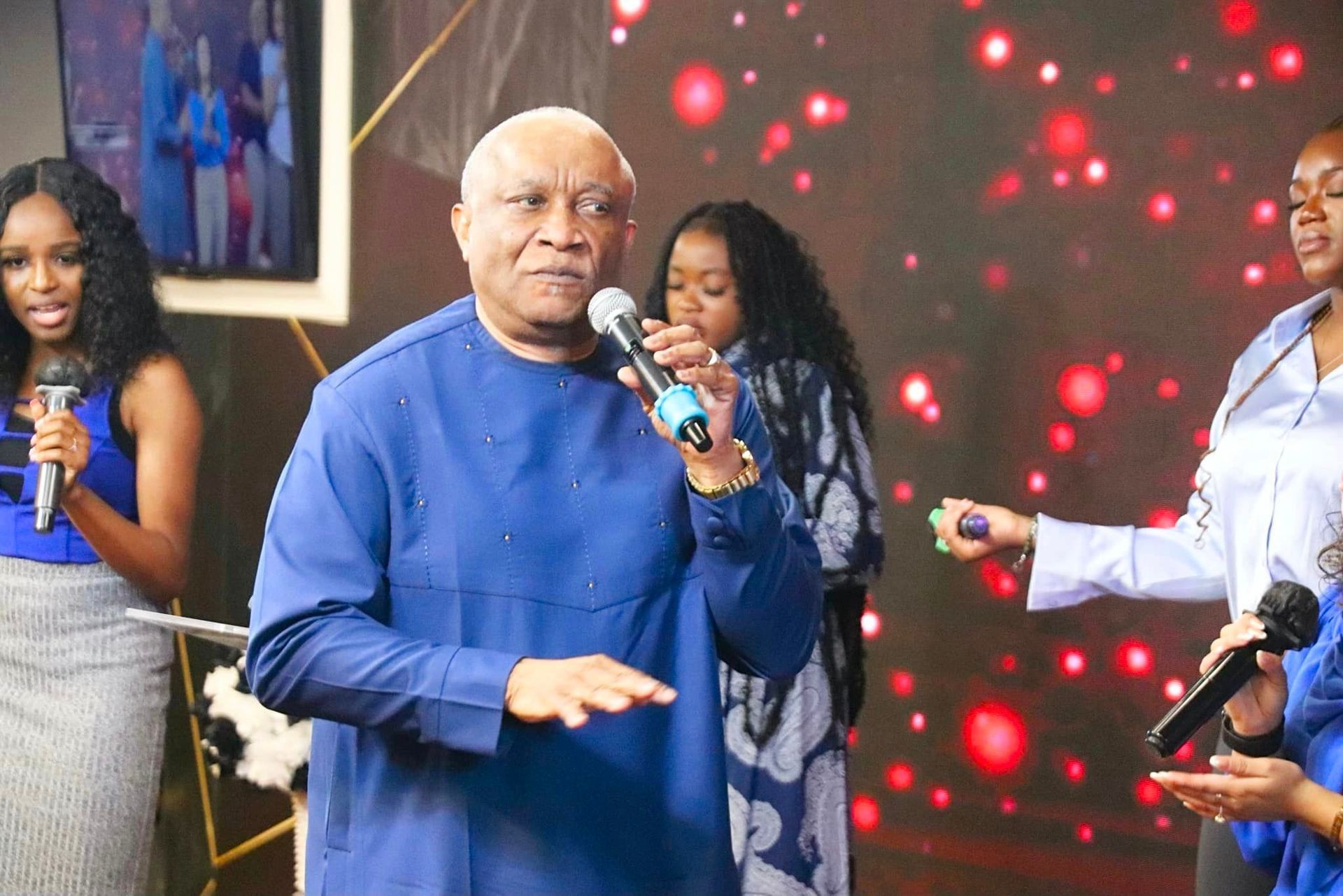 Man in blue shirt singing, surrounded by women with microphones, on a stage with a red backdrop.