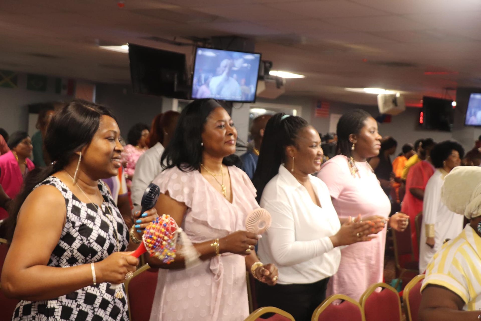 Women in a church service, clapping and singing. Some wear pink, holding maracas, and smiling.