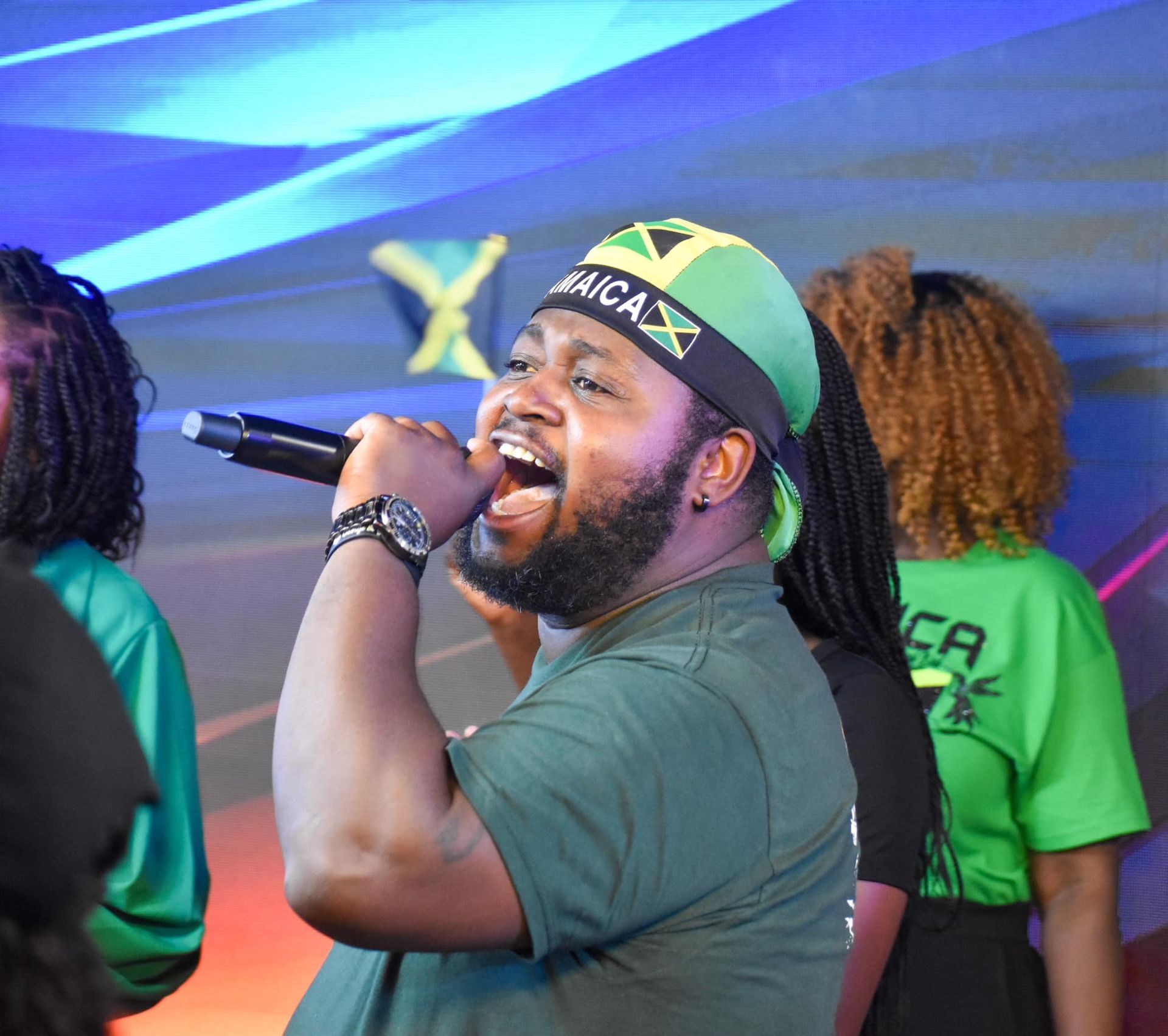 Man singing into a microphone wearing a Jamaican flag bandana and green shirt.