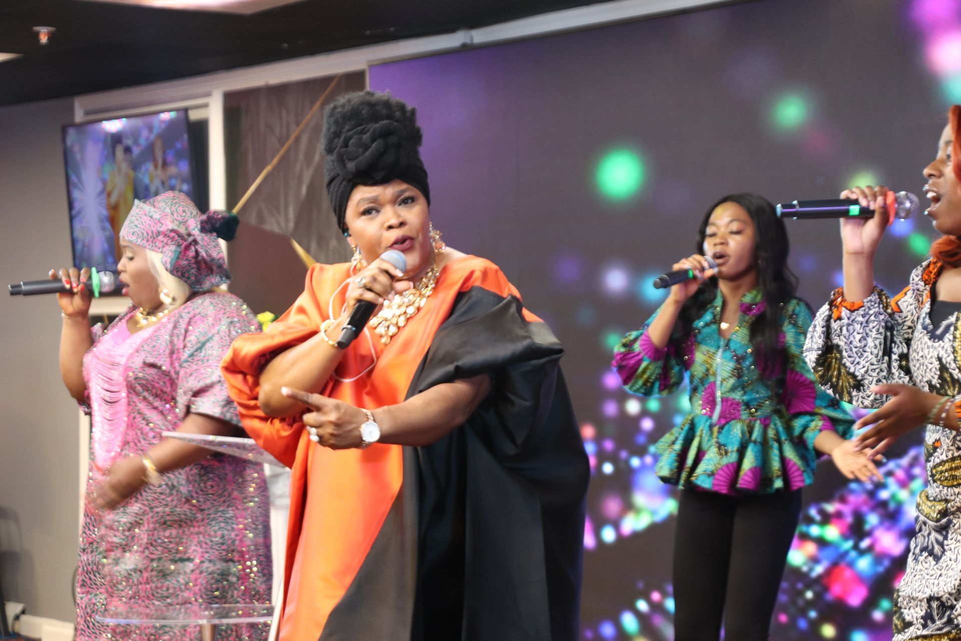 Women singing onstage, dressed in colorful African-inspired outfits. Stage lit with colorful lights.
