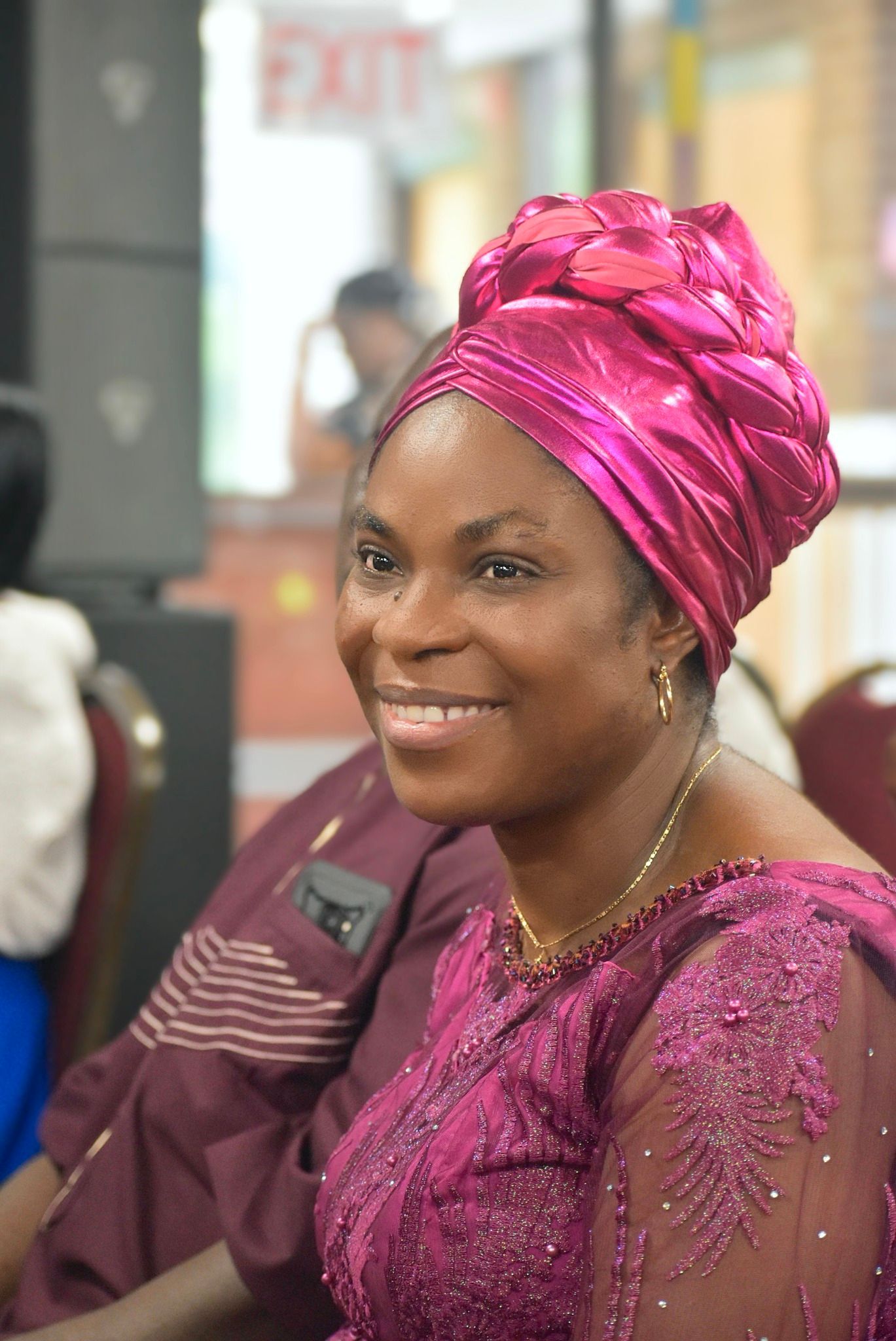 Woman wearing a pink headwrap and dress smiles, seated indoors.