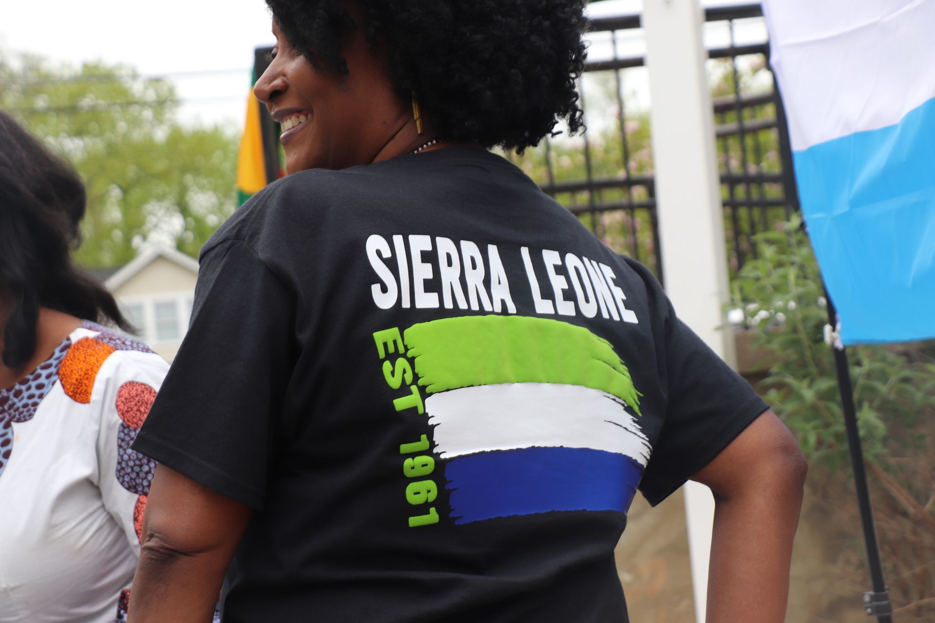 Woman wearing a black Sierra Leone shirt, smiling. The shirt has the Sierra Leone flag on the back, outdoors.