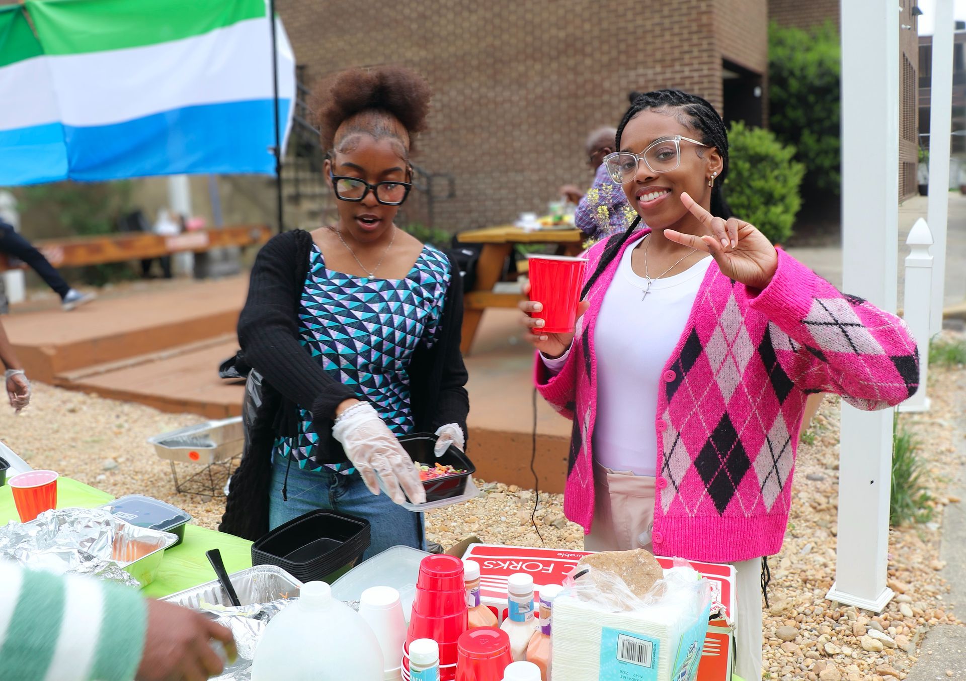 Two young women smiling at a food table with the Sierra Leone flag in the background.
