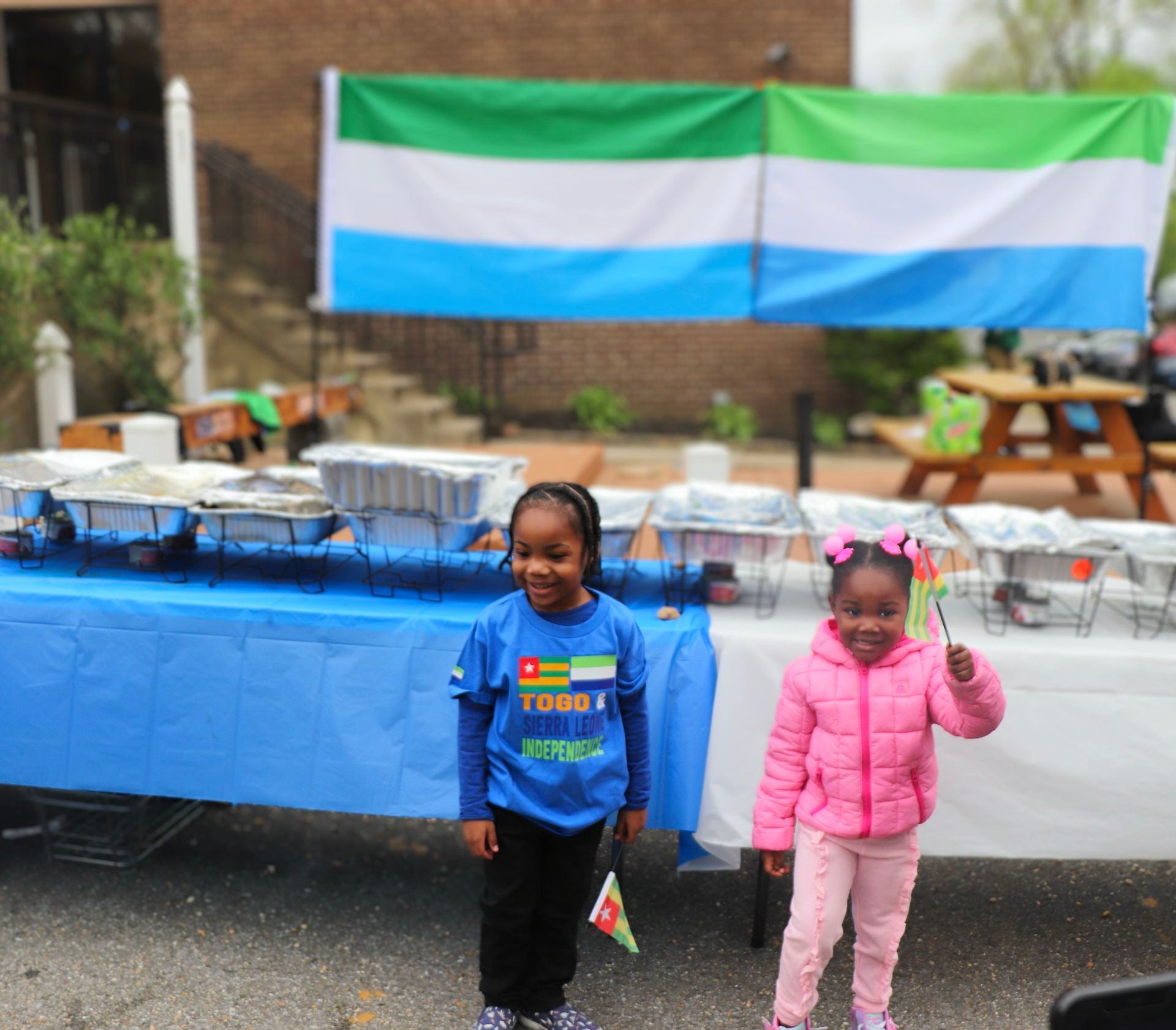 Two young girls in front of a table of food and a Sierra Leone flag. The girls hold flags.