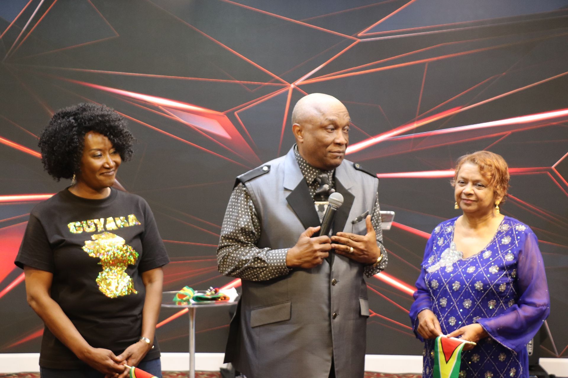 Three people standing on a stage; a man with a microphone speaks as two women beside him hold flags.