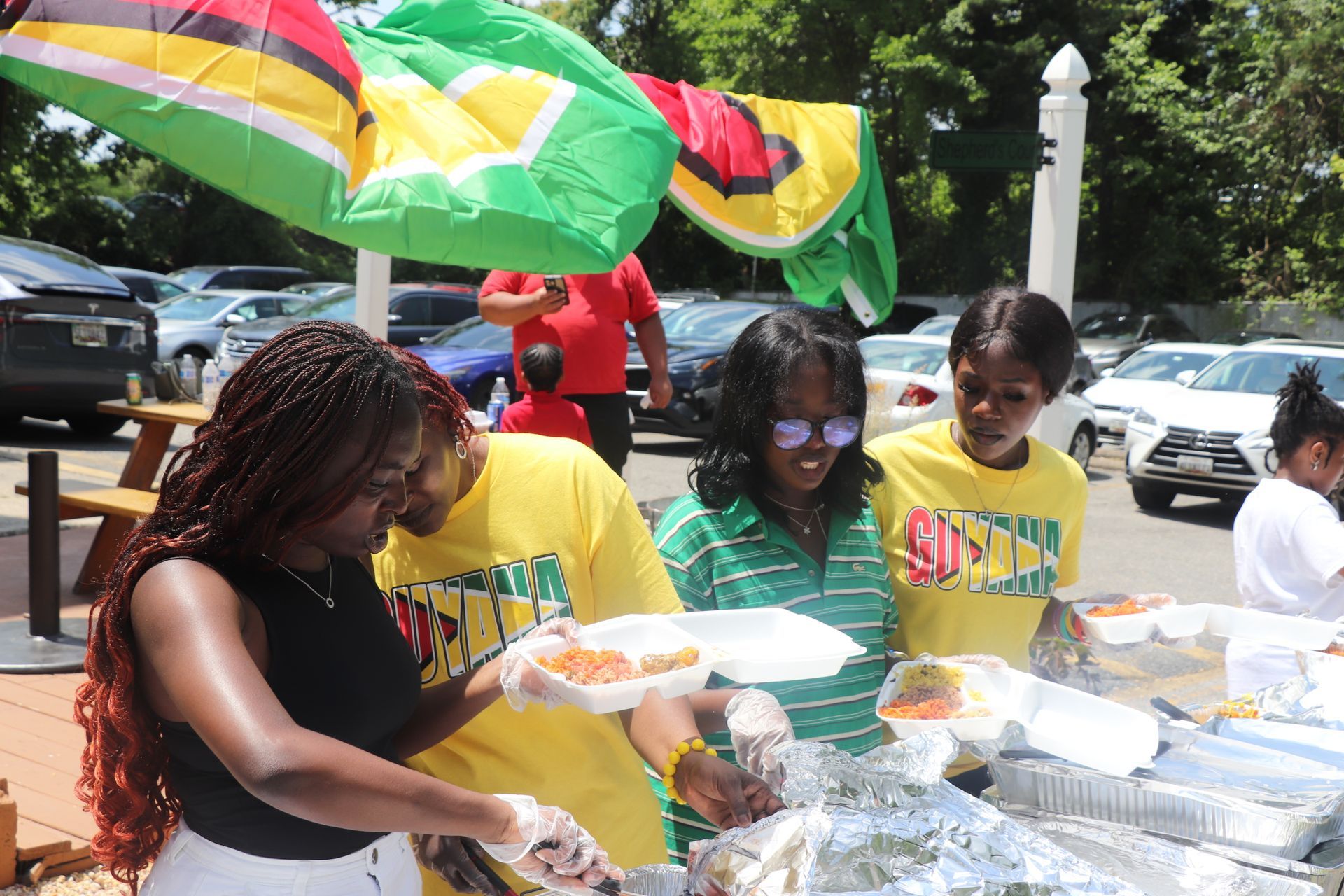 People serving food at a Guyanese festival. Colorful flags overhead.