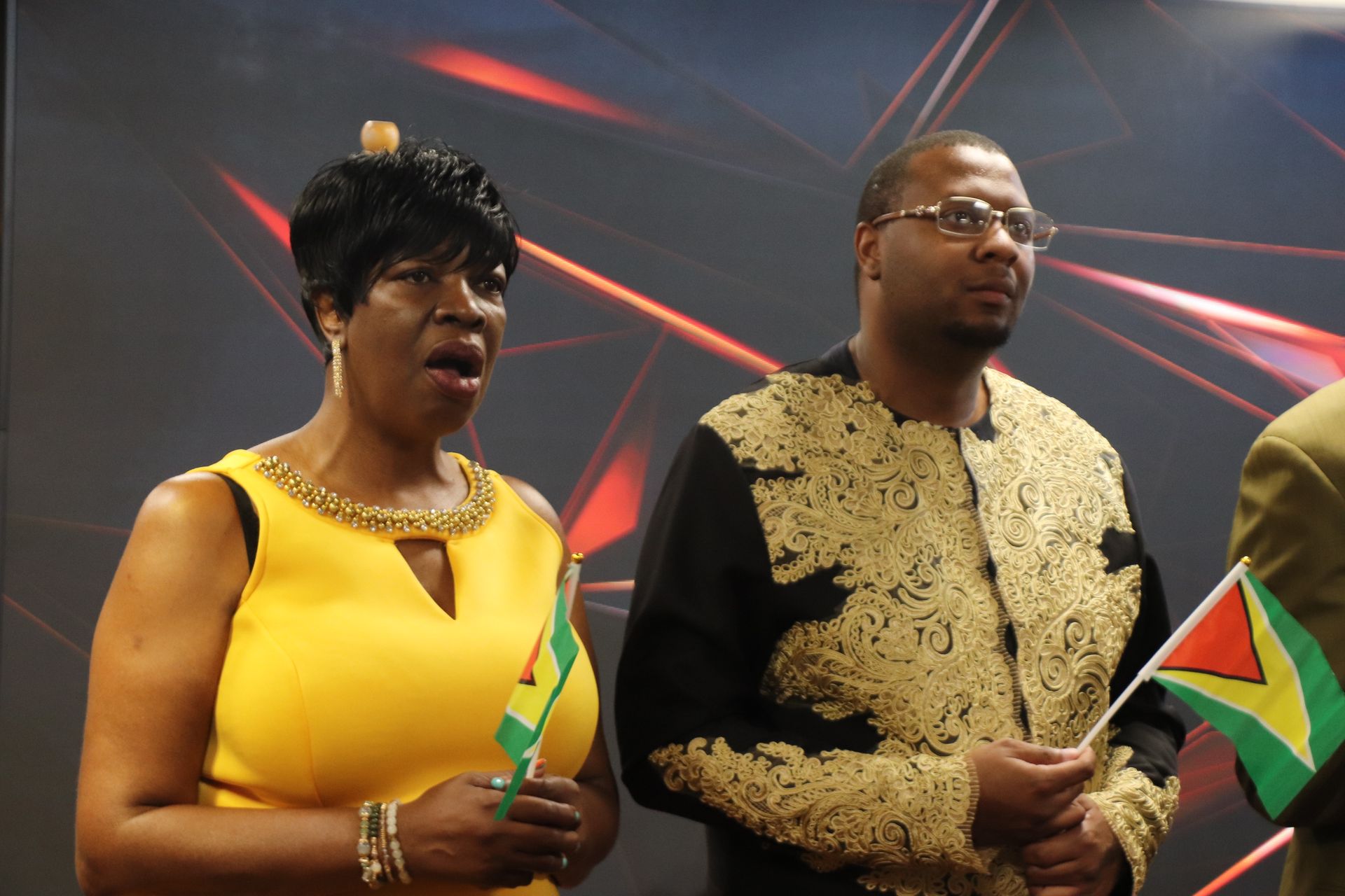 Two people, a woman in yellow and a man in black with gold, holding flags, singing.