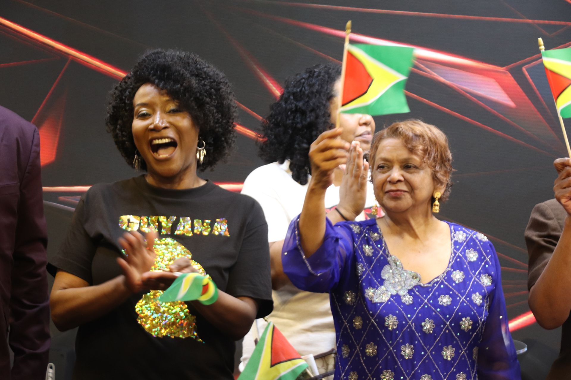People celebrating, waving Guyana flags. Woman in black shirt claps, others smile in a studio setting.