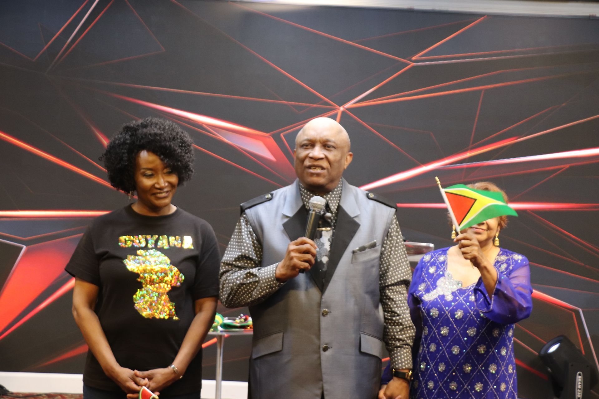 Three people stand on stage, a man speaking into a microphone, women on either side holding a Guyana flag and a T-shirt.