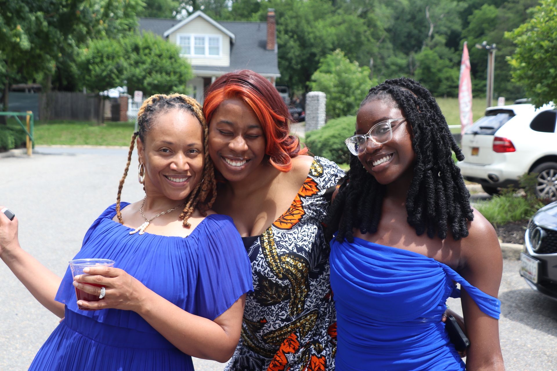 Three smiling women pose outdoors: one with braids, one with red hair, and one in a blue dress.